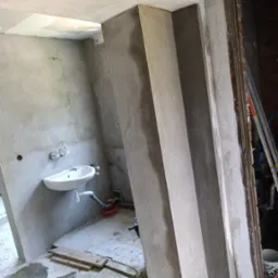 Wnętrze w trakcie prac wykończeniowych: otynkowane ściany, umywalka, widoczne ślady prac budowlanych. Fragment futryny drzwi i drabina w tle.