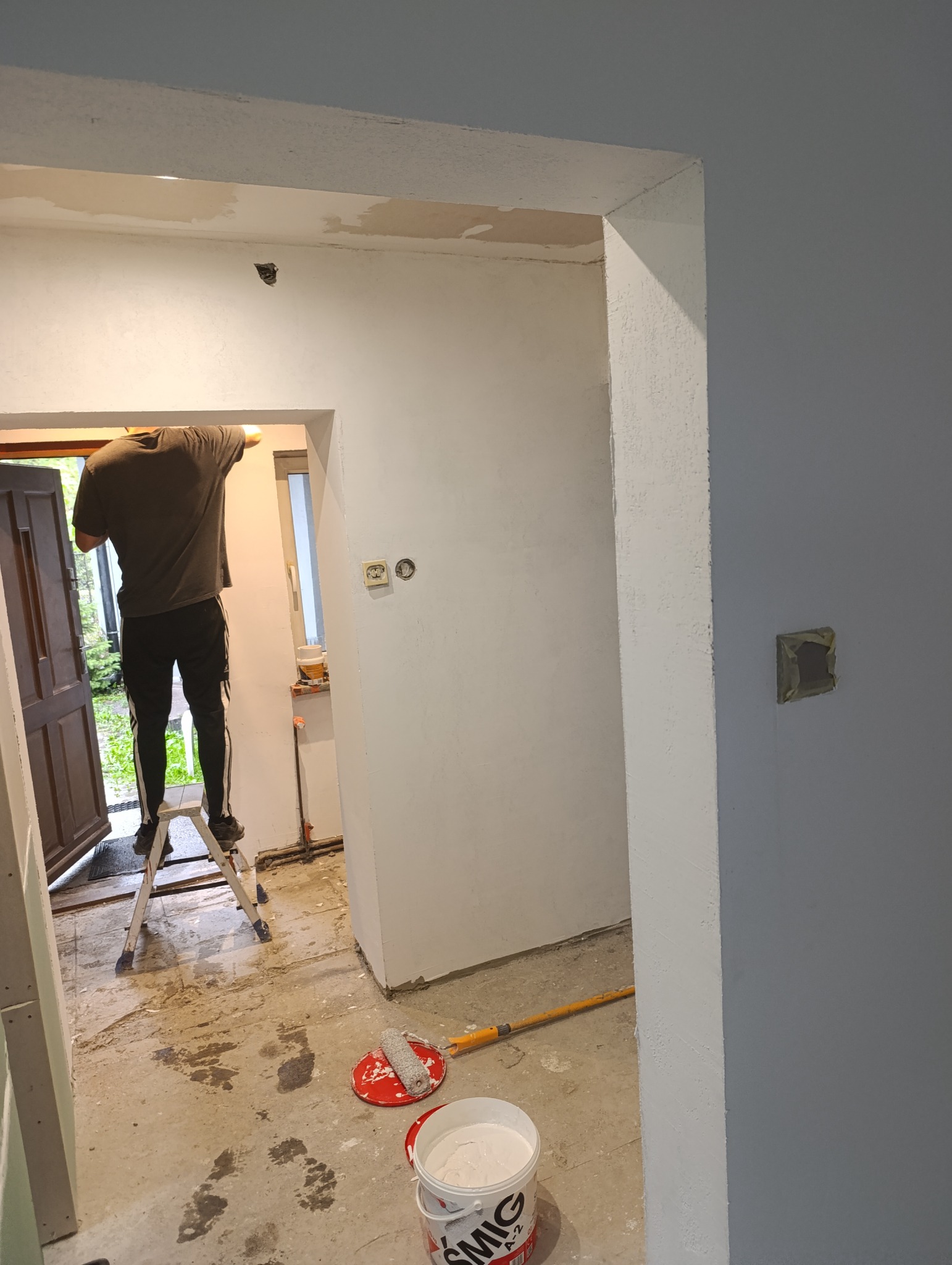 Prace wykończeniowe w Łasku: mężczyzna na drabinie maluje ścianę na biało, widoczne narzędzia malarskie i surowy stan podłogi.