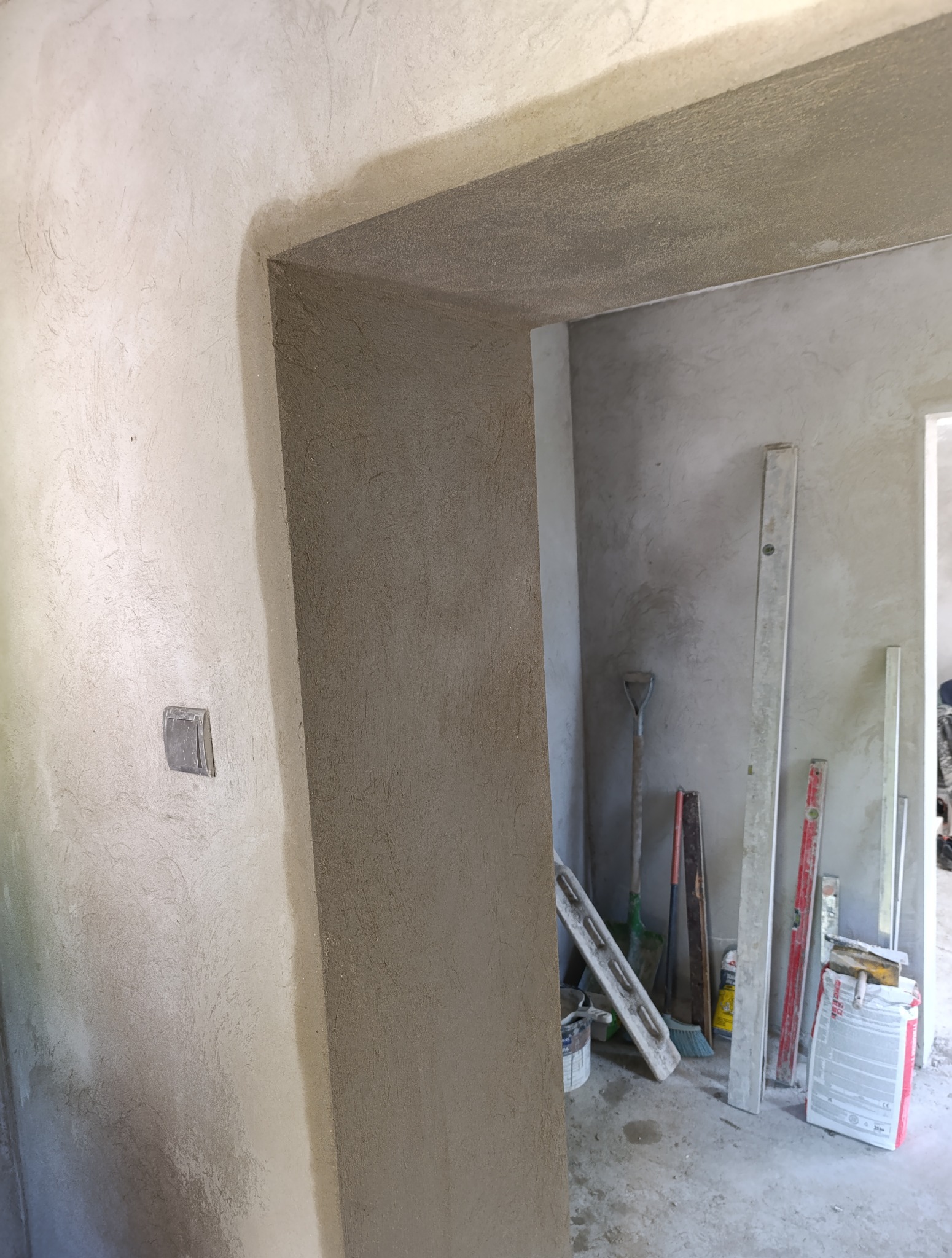 Otynkowane przejście drzwiowe z widocznymi narzędziami tynkarskimi w tle. Świeżo nałożony tynk o fakturze baranka. Widoczny włącznik światła na ścianie.