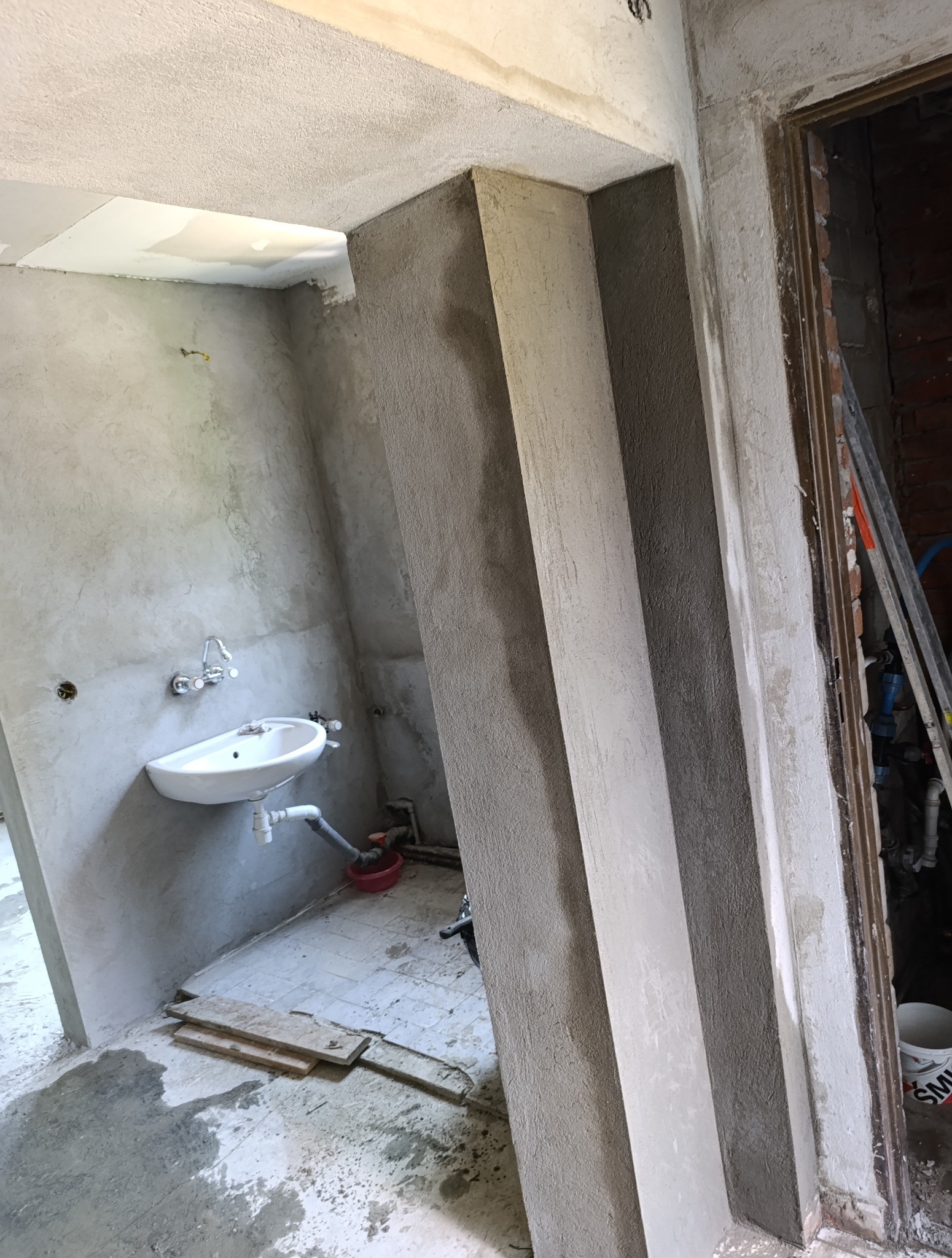 Wnętrze w trakcie prac wykończeniowych: otynkowane ściany, umywalka, widoczne ślady prac budowlanych. Fragment futryny drzwi i drabina w tle.