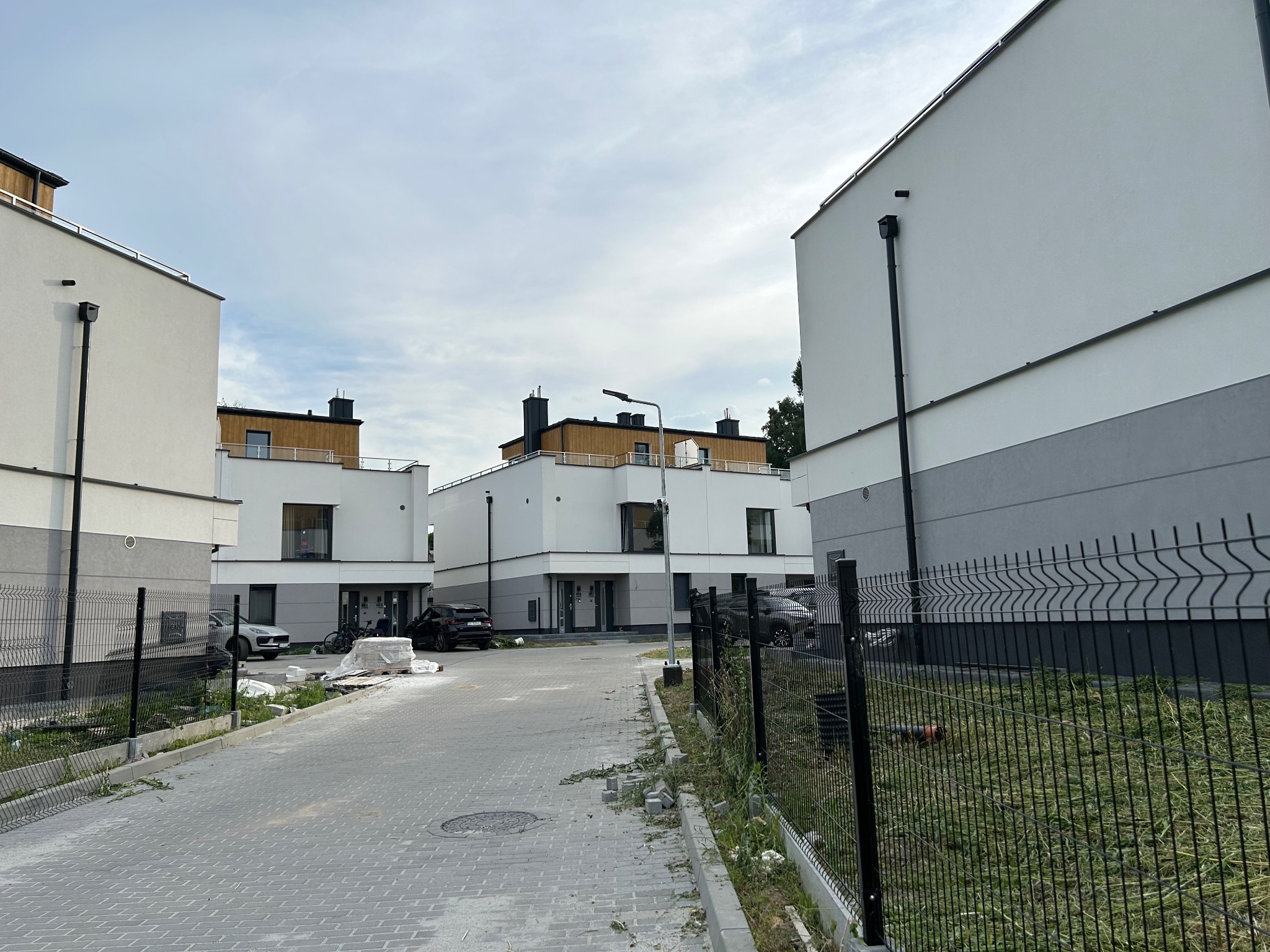 Nowoczesne osiedle z białymi domami i ciemnymi dachami, widok z poziomu ulicy z elementami montażu ogrodzenia panelowego.