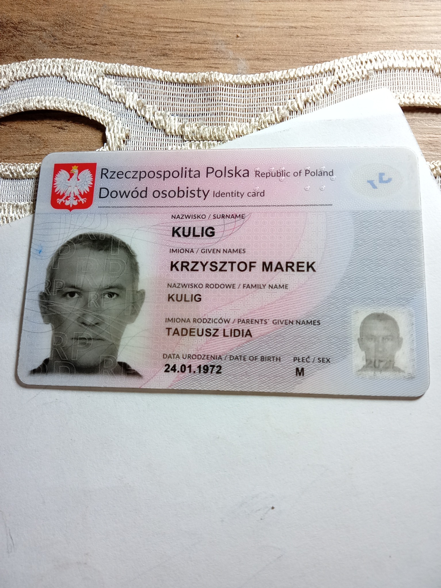 Dowód osobisty Rzeczypospolitej Polskiej na tle białej serwetki. Widoczne dane osobowe: nazwisko, imiona, imiona rodziców, data urodzenia, płeć oraz zdjęcie.