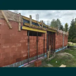 Budowa domu z czerwonej cegły: ściany w trakcie murowania, widoczne podpory, drewniane szalunki i izolacja fundamentów. Etap wznoszenia ścian.