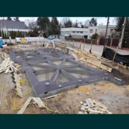 Zalane betonem fundamenty domu jednorodzinnego z wystającymi prętami zbrojeniowymi, widok z góry na plac budowy w słoneczny dzień.