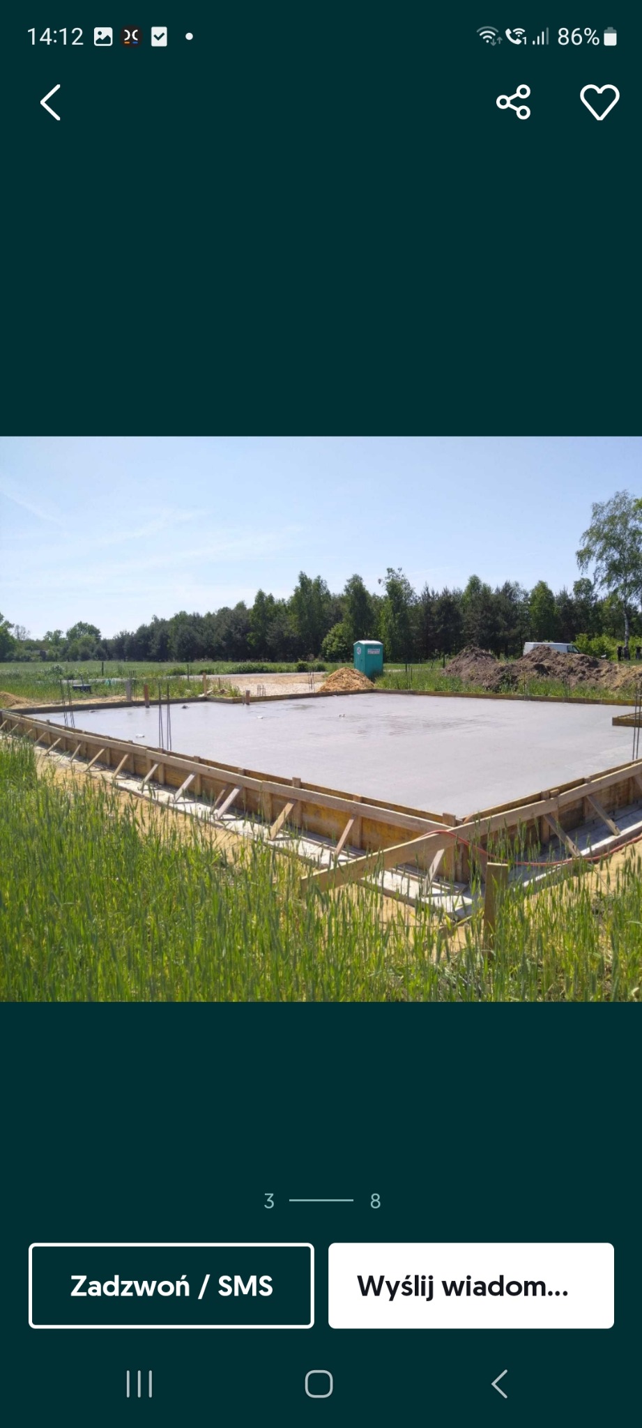 Wylany fundament pod budowę domu, drewniane szalunki, zbrojenie, widok z poziomu trawy, w tle drzewa i przenośna toaleta.