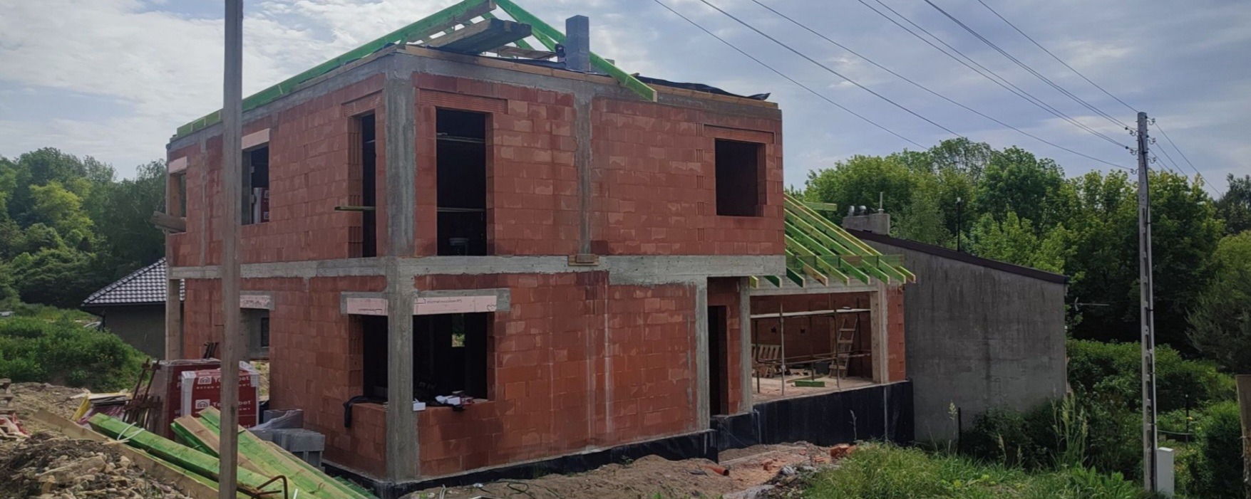 Dwukondygnacyjny dom w trakcie budowy, ściany z czerwonej cegły, widoczne otwory okienne i zarys dachu z zielonymi elementami. W tle zieleń i fragment sąsiedniego budynku.