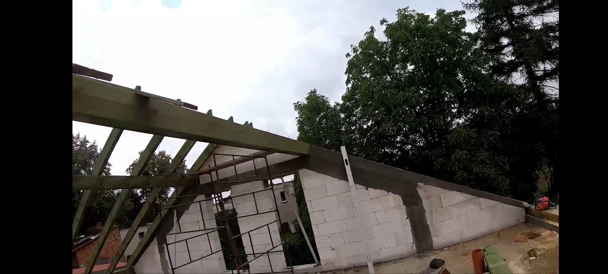Budowa domu: wzniesione ściany z betonu komórkowego, widoczna konstrukcja więźby dachowej. Na ścianie przymocowana poziomica. W tle drzewa i pochmurne niebo.