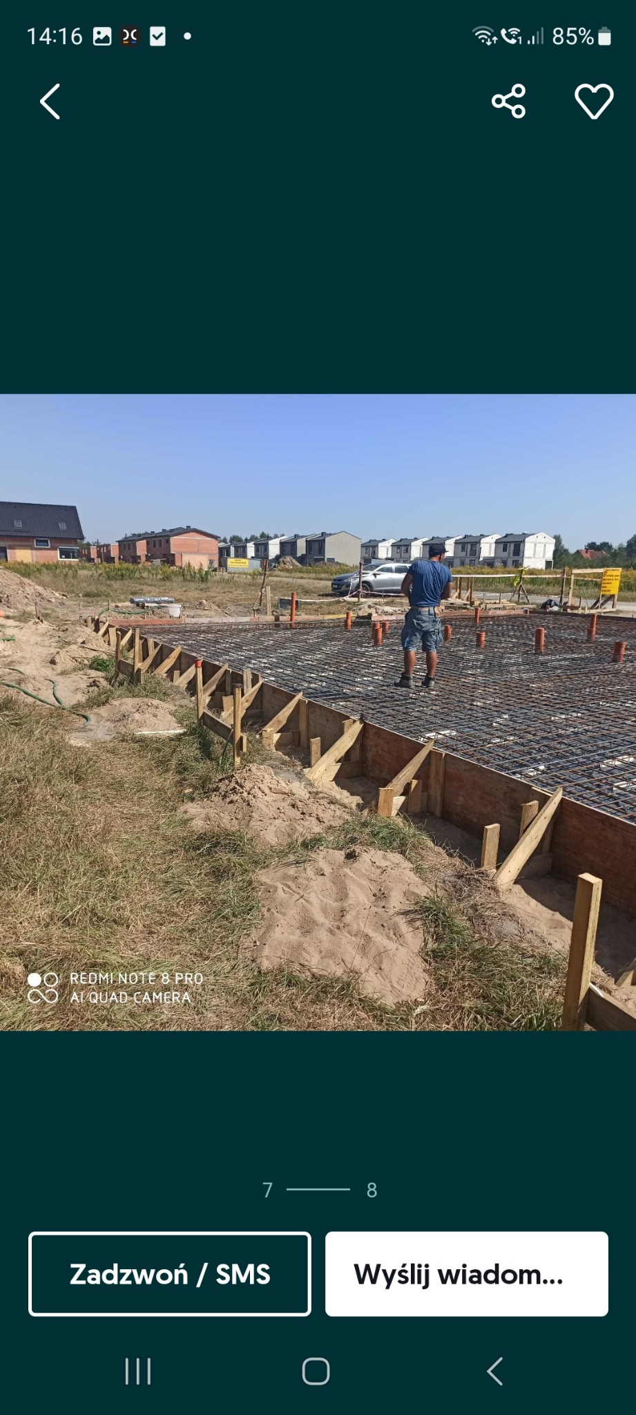 Zbrojenie fundamentów domu jednorodzinnego z drewnianym szalunkiem i pionowymi rurami. W tle widoczne domy w zabudowie szeregowej i pracownik budowlany.