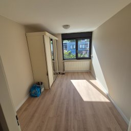 KADET STYL I PRECYZJA - Jasne, puste pomieszczenie po remoncie z drewnianą podłogą, beżowymi ścianami i białymi drzwiami szafy. Promień słońca wpada przez okno. Widok na budynki.