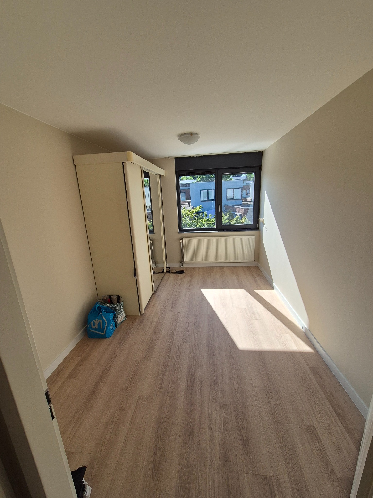 Jasne, puste pomieszczenie po remoncie z drewnianą podłogą, beżowymi ścianami i białymi drzwiami szafy. Promień słońca wpada przez okno. Widok na budynki.