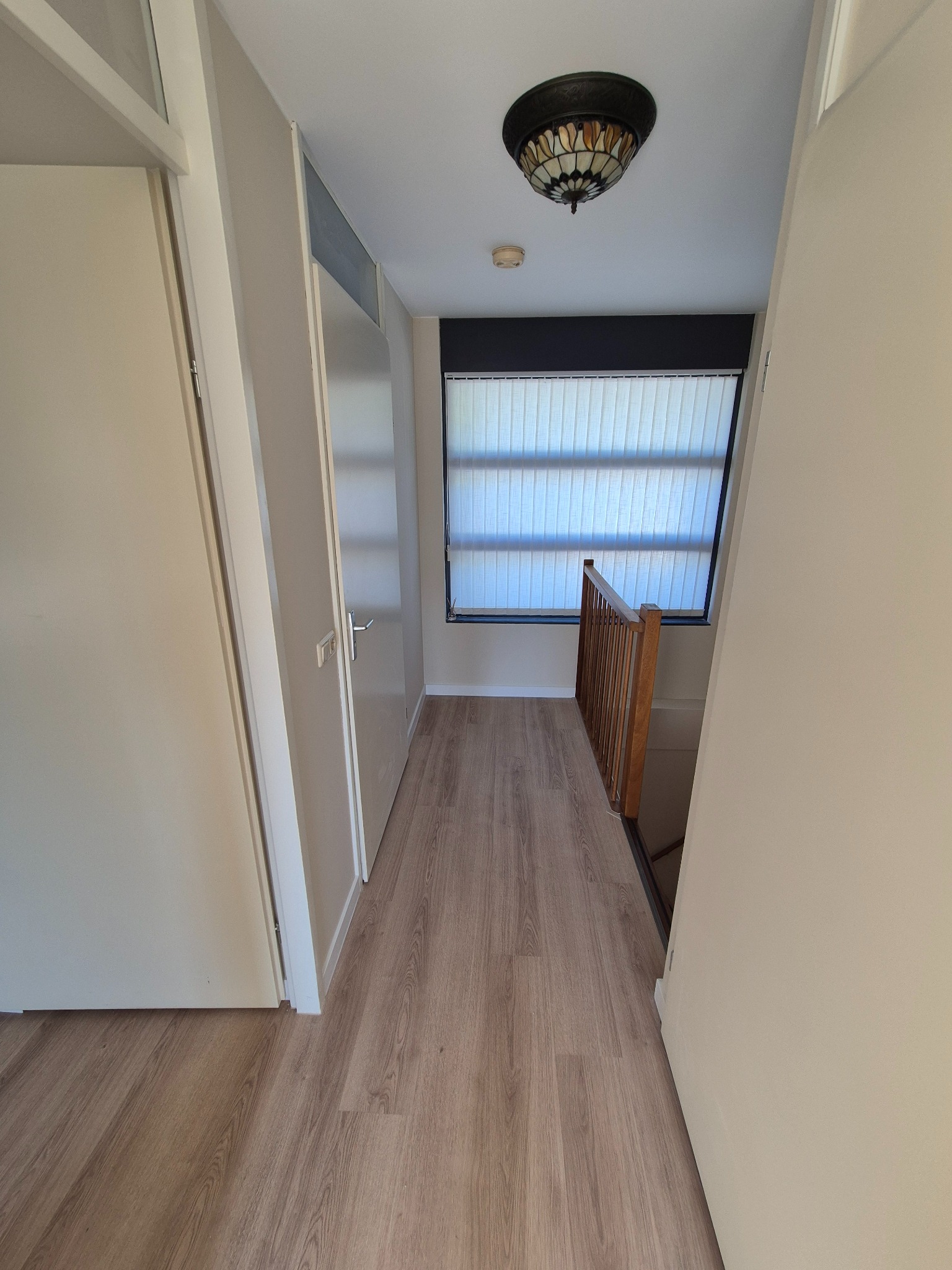 Korytarz z drewnianą podłogą i białymi ścianami. Widoczne drzwi, okno z żaluzjami i ozdobna lampa sufitowa. Neutralna kolorystyka i nowoczesny wystrój.