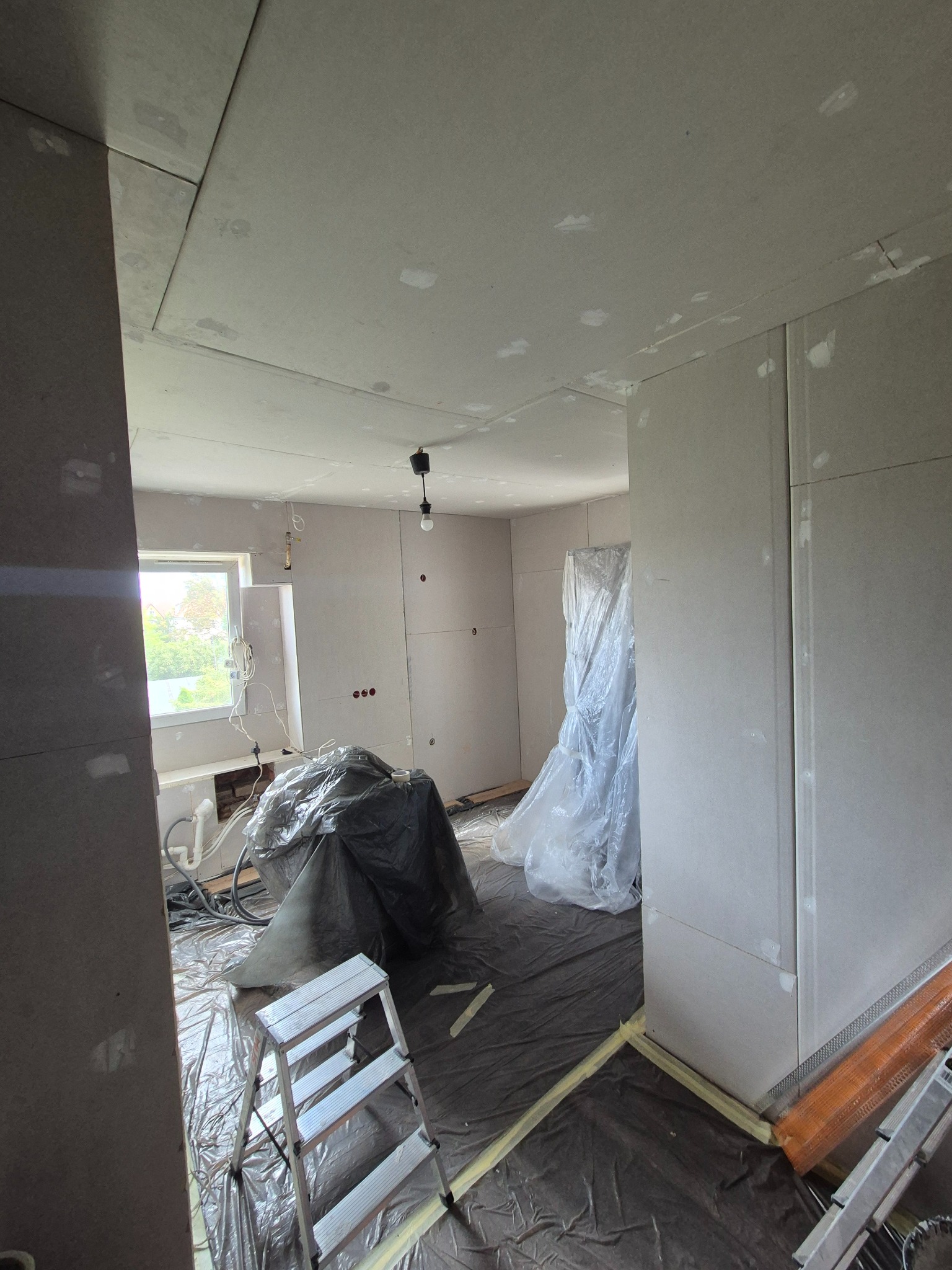 Pokój w trakcie montażu płyt kartonowo-gipsowych, widoczne płyty na ścianach i suficie, zakryte folią elementy, drabina. Prace wykończeniowe w jasnym pomieszczeniu.