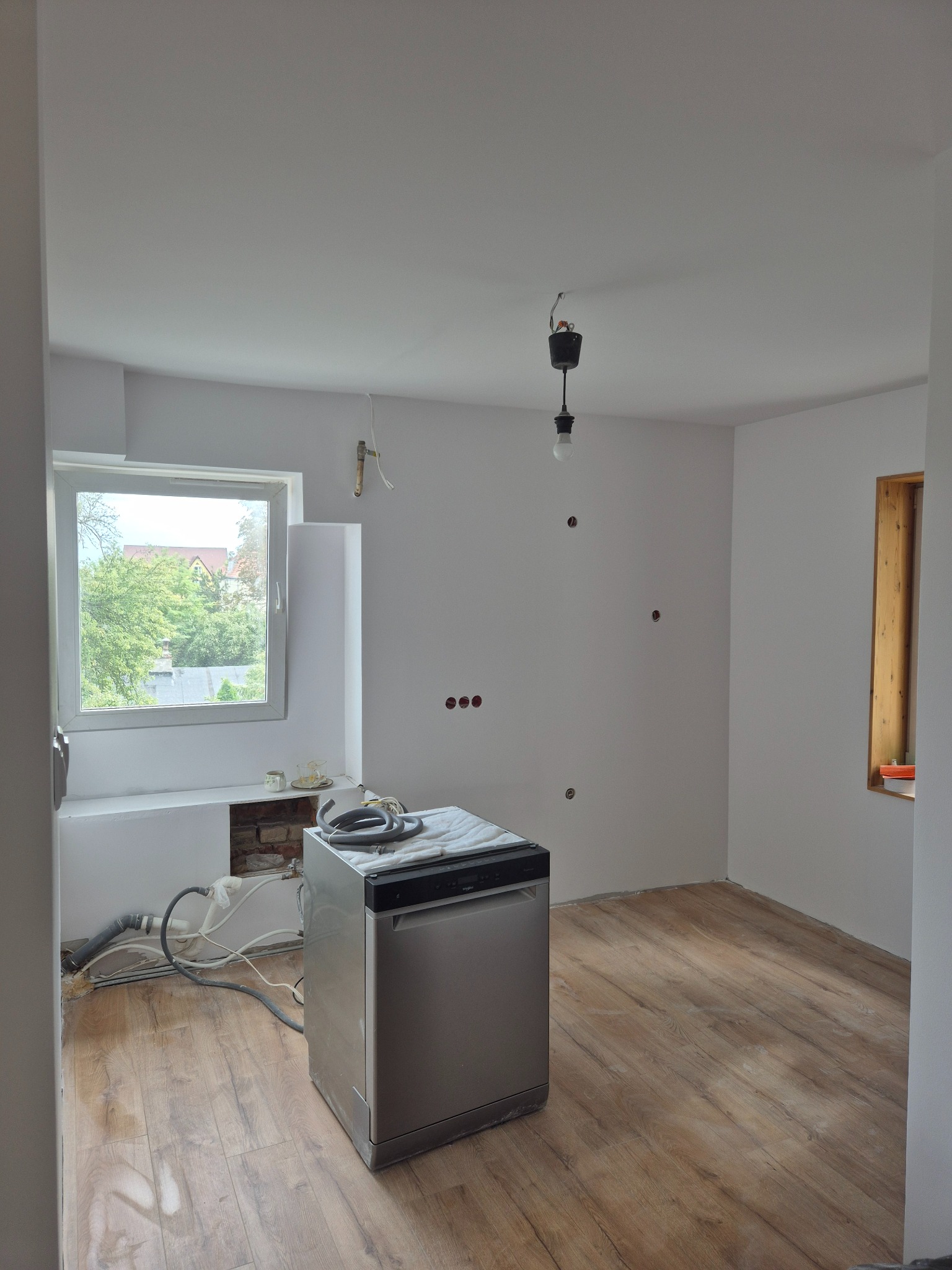 Wnętrze pokoju po remoncie: świeżo pomalowane ściany na biało, drewniana podłoga, okno z widokiem na zieleń, pralka. Widoczne ślady po instalacjach elektrycznych.