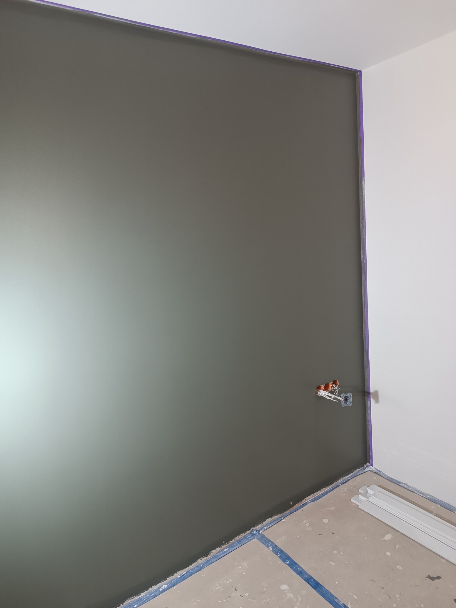 Ściana pomalowana na szaro, zabezpieczona taśmą malarską, z wystającą puszką elektryczną i listwami przypodłogowymi. Widok na róg pokoju.