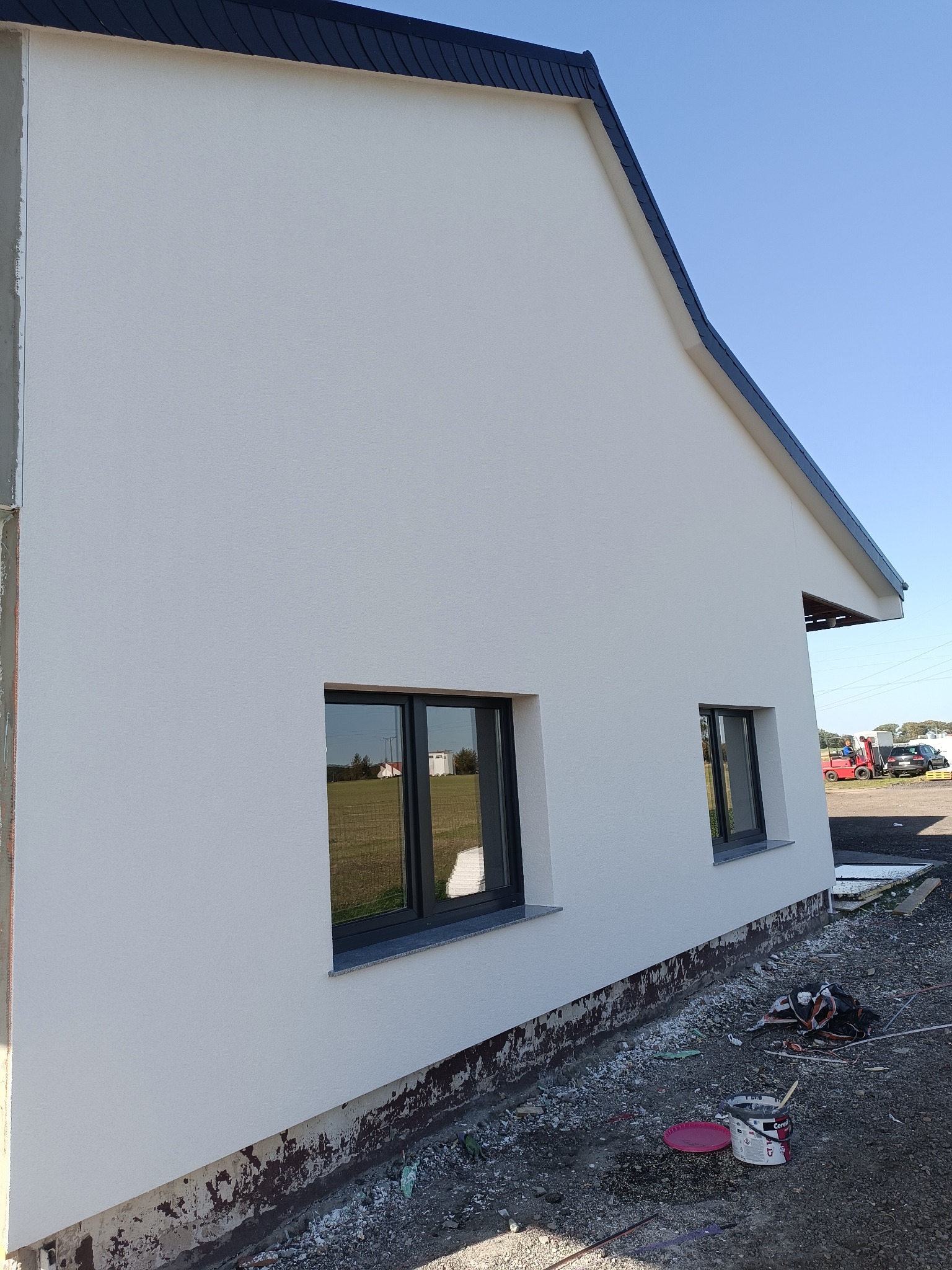 Boczny widok domu po nałożeniu białego tynku, z czarnymi oknami i dachem. Widoczne ślady po pracach wykończeniowych i kubeł z farbą.