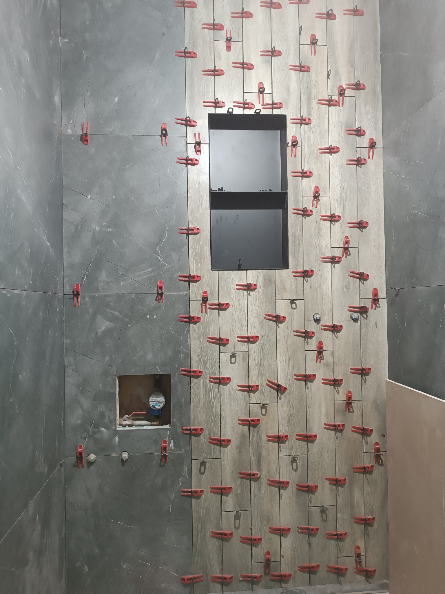 Montaż płytek imitujących drewno w łazience z użyciem czerwonych klipsów poziomujących. Widoczna wnęka i otwór na licznik.