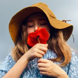 Dziewczyna w słomkowym kapeluszu z zamkniętymi oczami wącha czerwonego maku na tle pochmurnego nieba. Ubrana w bluzkę w etniczne wzory.