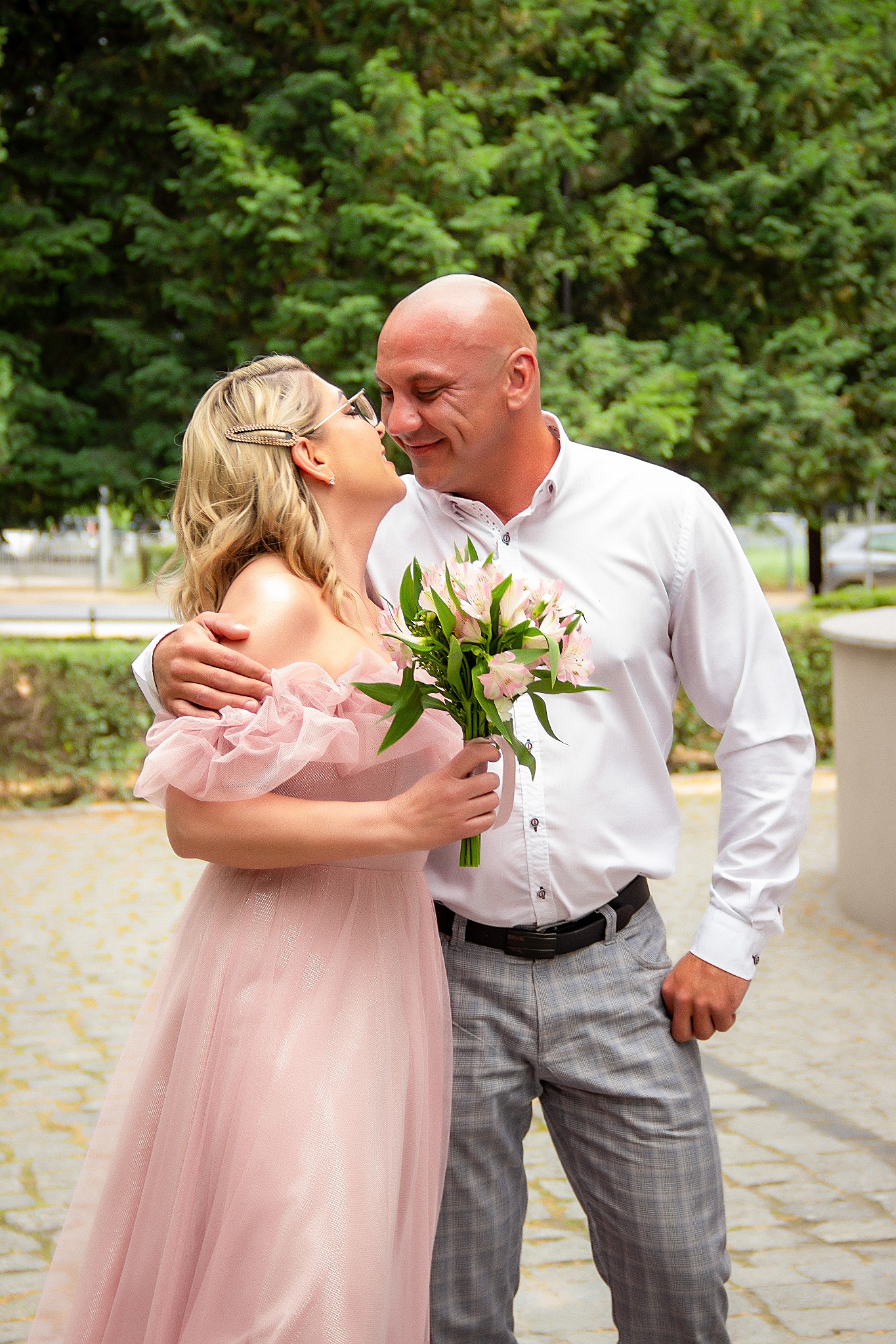 Uśmiechnięta para w objęciach na tle zieleni. Kobieta w pudrowej sukience trzyma bukiet, mężczyzna w białej koszuli i szarych spodniach. Romantyczny moment.