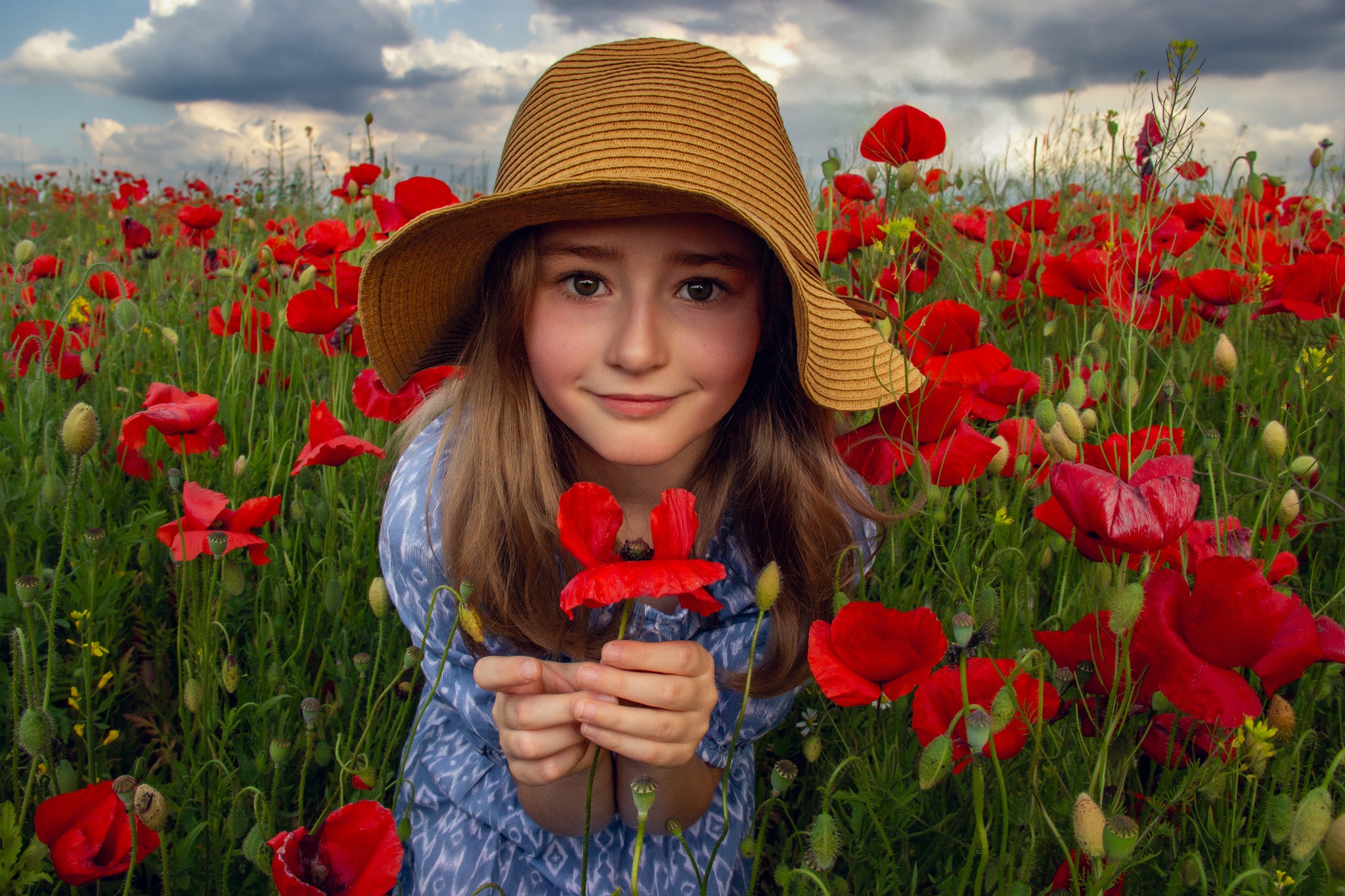 Uśmiechnięta dziewczynka w słomkowym kapeluszu trzyma mak na tle kwitnącego pola maków. Szeroki kadr, naturalne światło, głębia ostrości.
