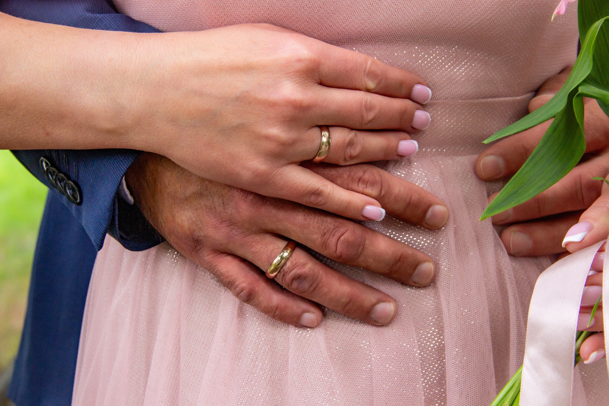 Dłonie pary z obrączkami na tle różowej sukienki i bukietu, zbliżenie na detale ślubne, subtelne oświetlenie, kadrowanie na dłonie.