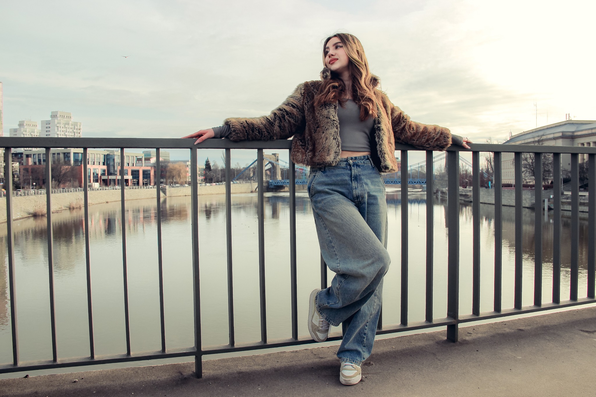 Kobieta w futrze opiera się o barierkę mostu, spoglądając w bok. W tle woda, budynki i most. Ujęcie z dołu podkreśla perspektywę i sylwetkę modelki.