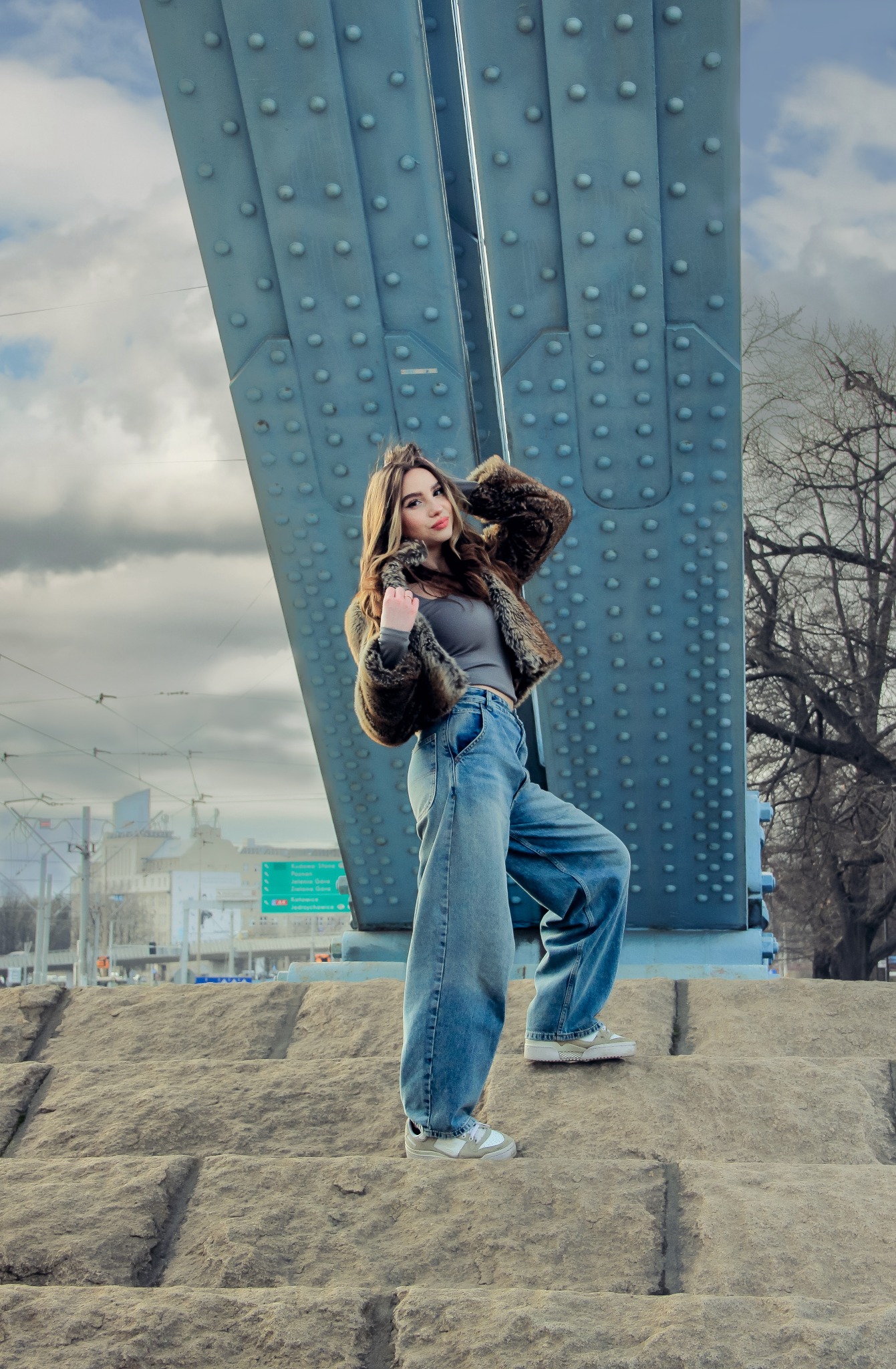 Kobieta w futrze i szerokich jeansach pozuje na tle stalowej konstrukcji mostu, z widokiem na miasto w tle. Ujęcie z dołu, podkreślające architekturę i styl.
