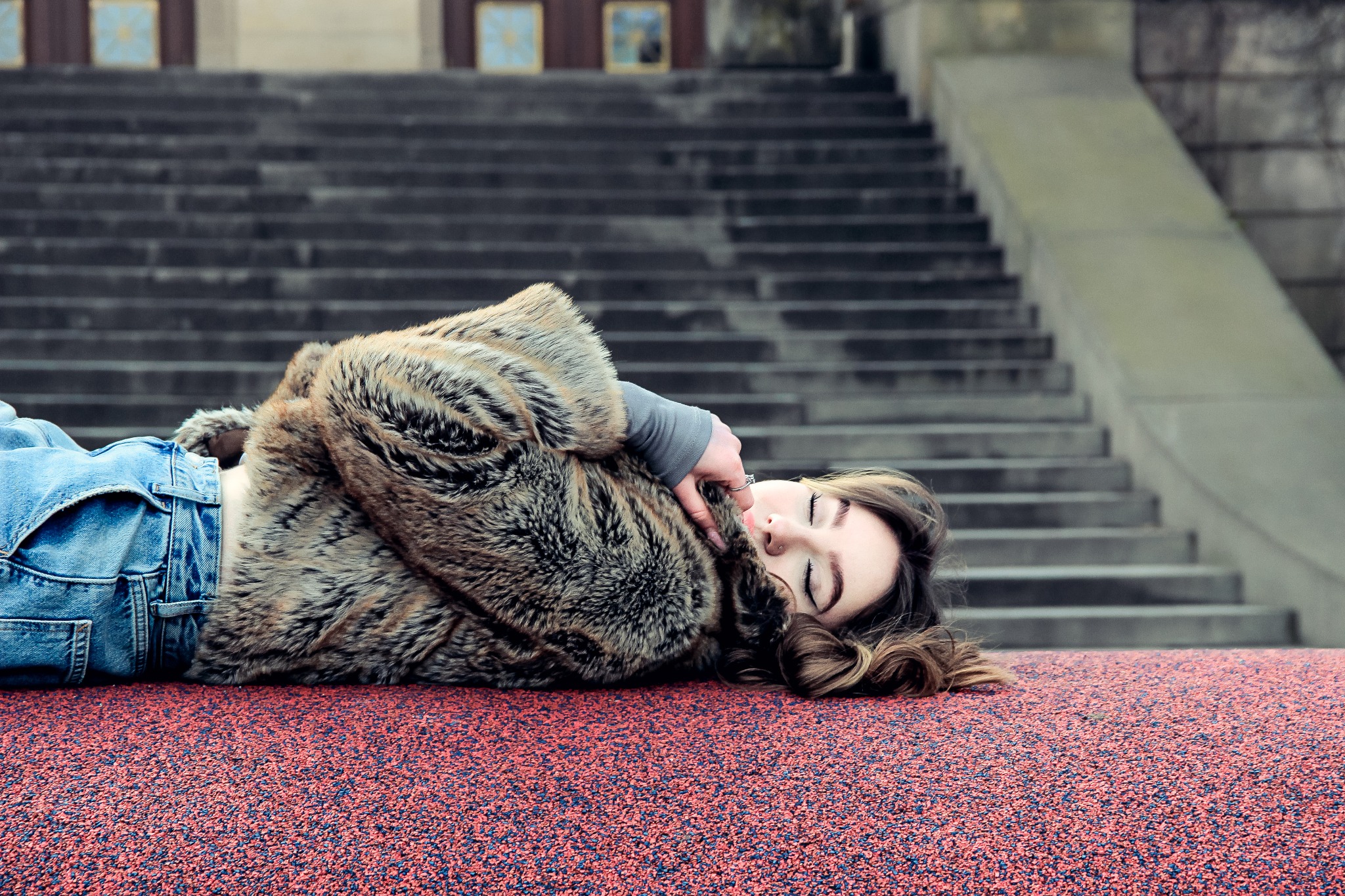 Kobieta w futrze leży na czerwonej powierzchni, tłem są schody. Ujęcie z góry, zamknięte oczy, delikatny uśmiech. Kadrowanie od pasa w górę. Naturalne światło.