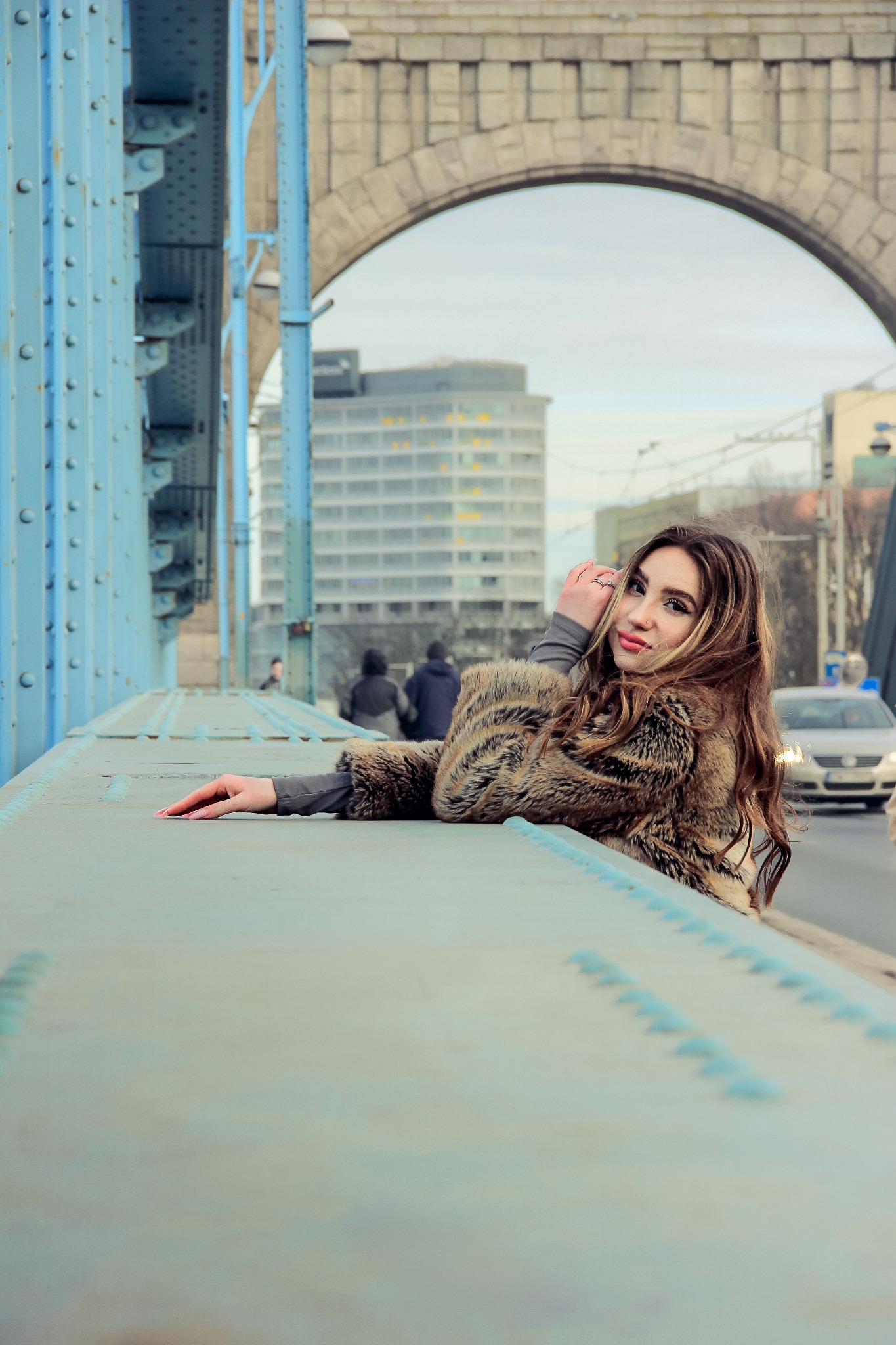 Kobieta w futrze opiera się o barierę na moście z niebieską konstrukcją. W tle budynek i ruch uliczny. Ujęcie z niskiej perspektywy, podkreślające architekturę.