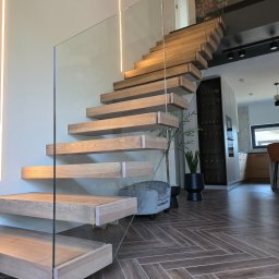 MAGIC FISH SPÓŁKA Z OGRANICZONĄ ODPOWIEDZIALNOŚCIĄ - Nowoczesne schody drewniane z minimalistyczną szklaną balustradą w jasnym wnętrzu z podświetleniem LED i parkietem w jodełkę. Widok na salon i kuchnię.
