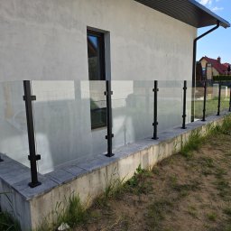 MAGIC FISH SPÓŁKA Z OGRANICZONĄ ODPOWIEDZIALNOŚCIĄ - Szklana balustrada z czarnymi słupkami, zamontowana na betonowej podmurówce, przylegająca do ściany budynku z oknem. Widok z zewnątrz, trawa i piasek w tle.