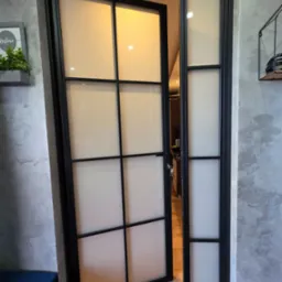 Otwarta, loftowa brama wewnętrzna z czarnymi profilami i mlecznym szkłem, widoczna z boku na tle szarej ściany i fragmentu podłogi.