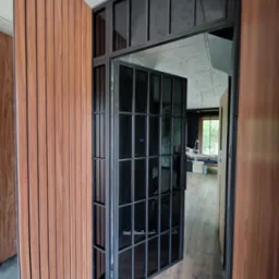 Nowoczesne, czarne, metalowe drzwi z szybami w loftowym stylu, otwarte do wnętrza. Obok widoczna drewniana okładzina ścienna w ciepłym odcieniu.