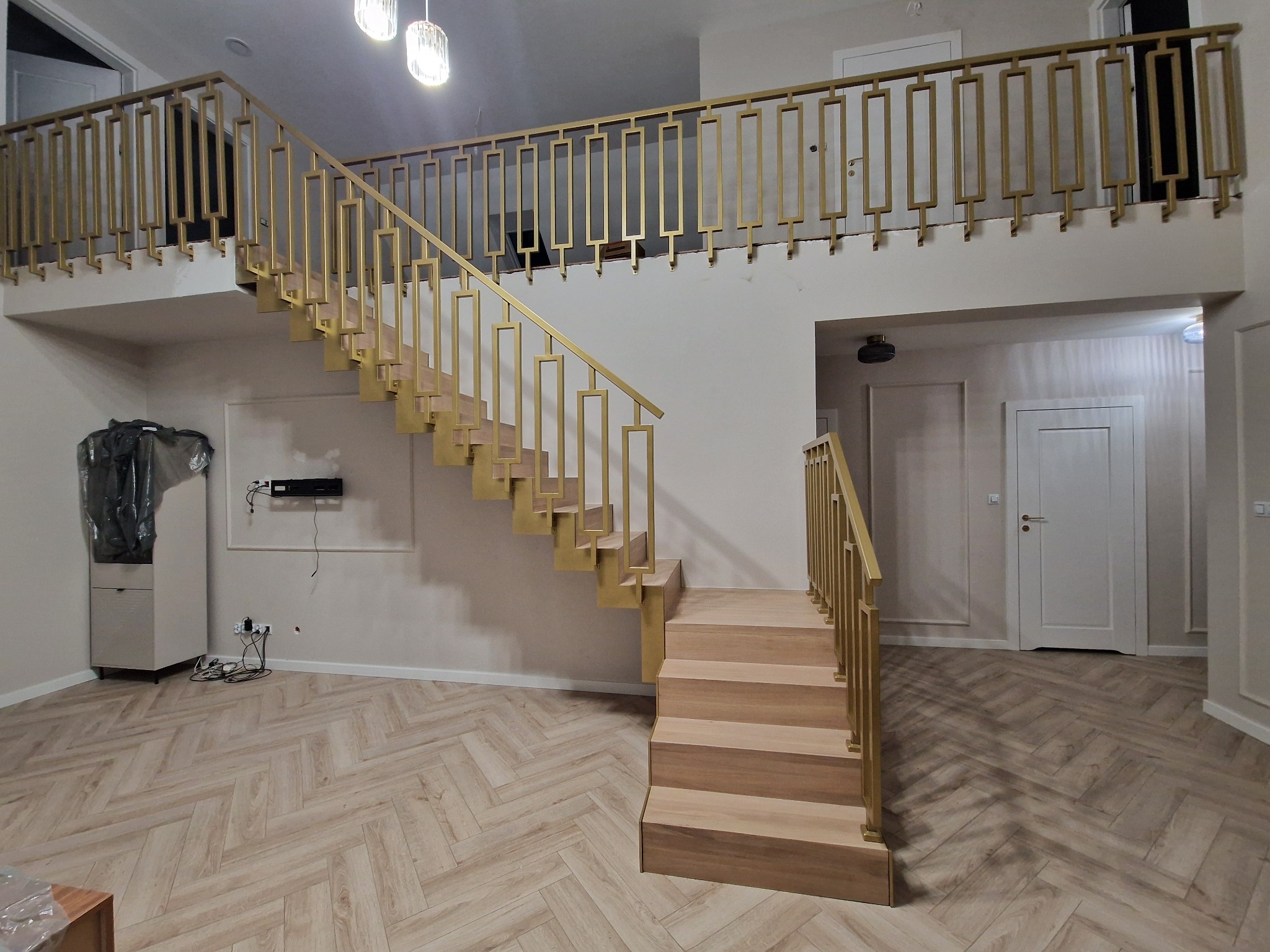 Nowoczesne schody drewniane z metalową, złocistą balustradą w dwupoziomowym wnętrzu. Widok na balustradę na antresoli i parkiet ułożony w jodełkę.