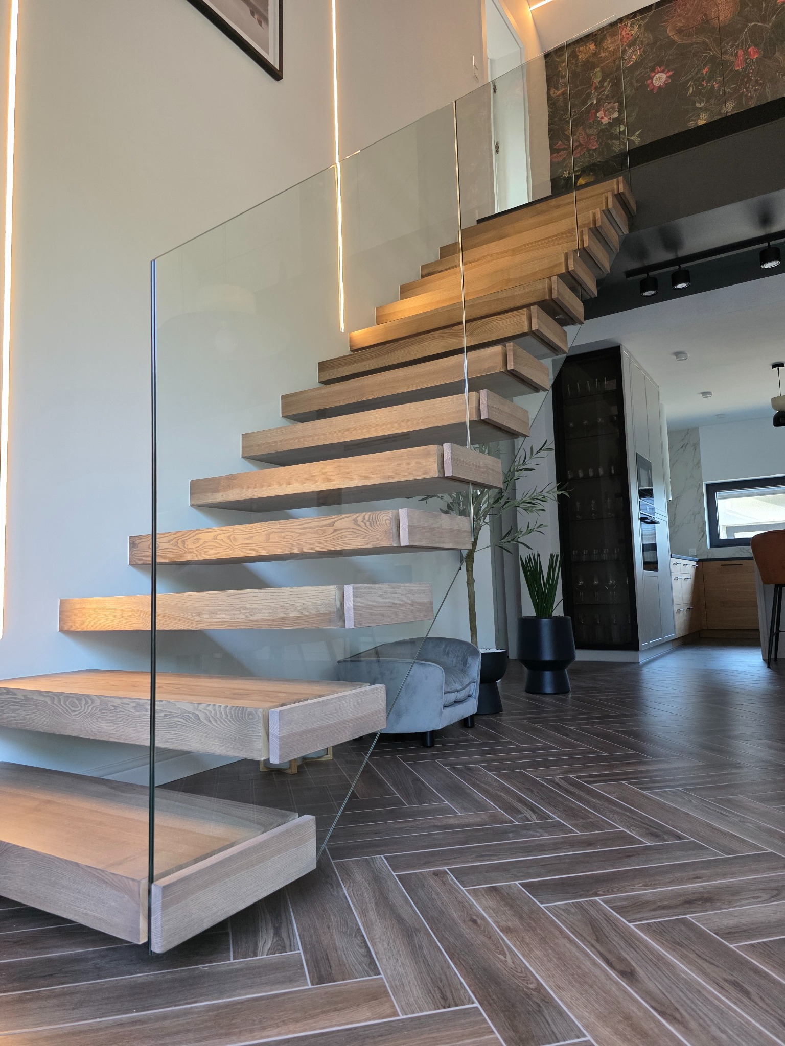 Nowoczesne schody drewniane z minimalistyczną szklaną balustradą w jasnym wnętrzu z podświetleniem LED i parkietem w jodełkę. Widok na salon i kuchnię.