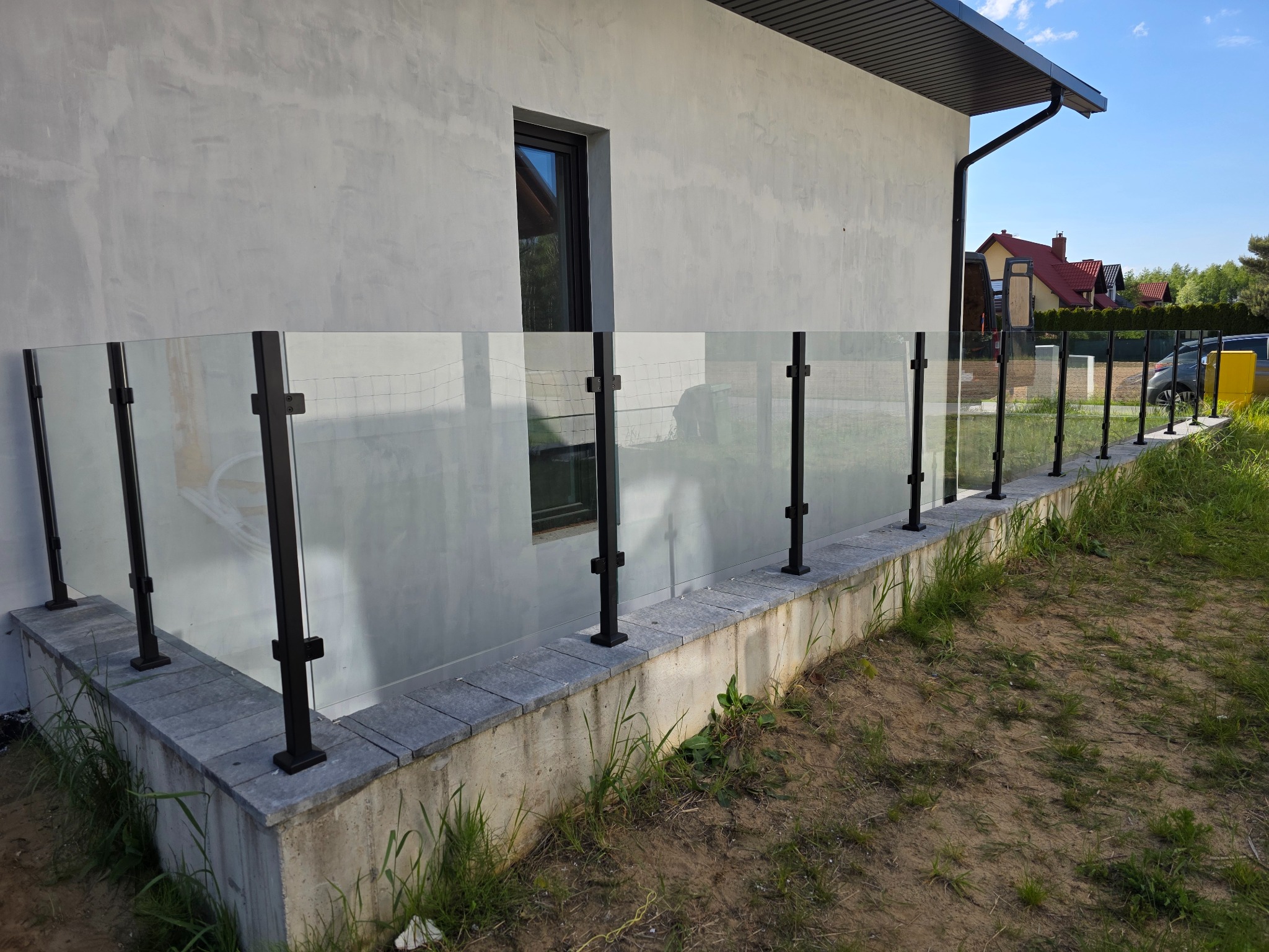 Szklana balustrada z czarnymi słupkami, zamontowana na betonowej podmurówce, przylegająca do ściany budynku z oknem. Widok z zewnątrz, trawa i piasek w tle.