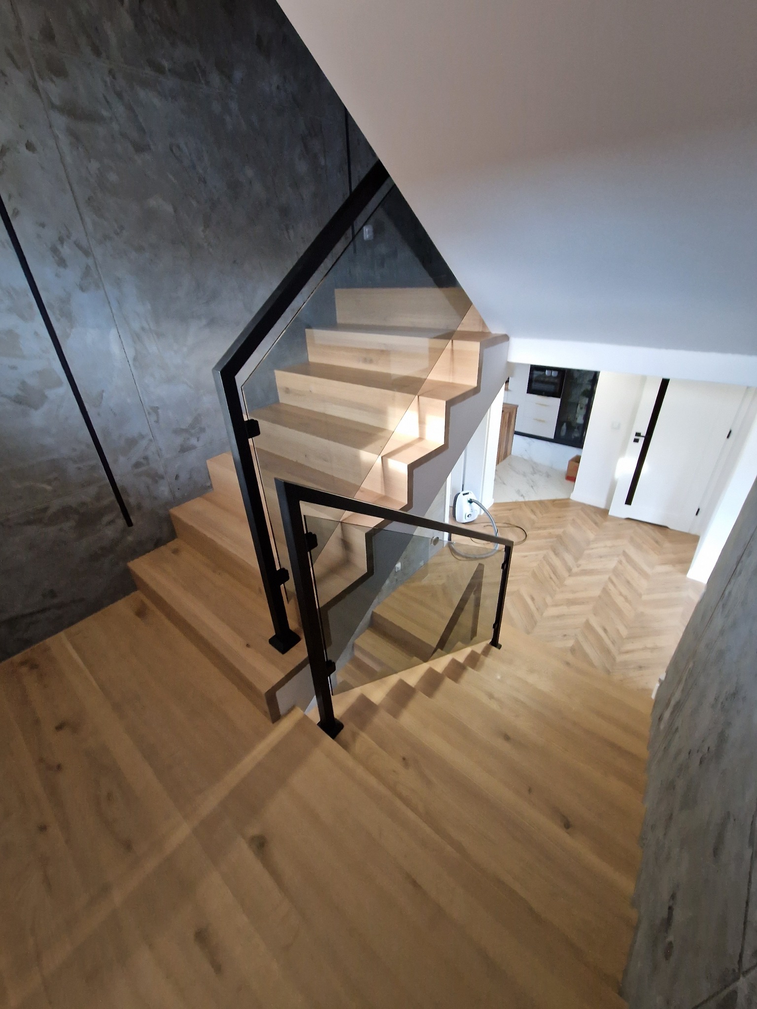 Drewniane schody z balustradą ze szkła i czarnymi elementami metalowymi, widok z góry, parkiet w jodełkę, szara ściana z efektem betonu, nowoczesny design.