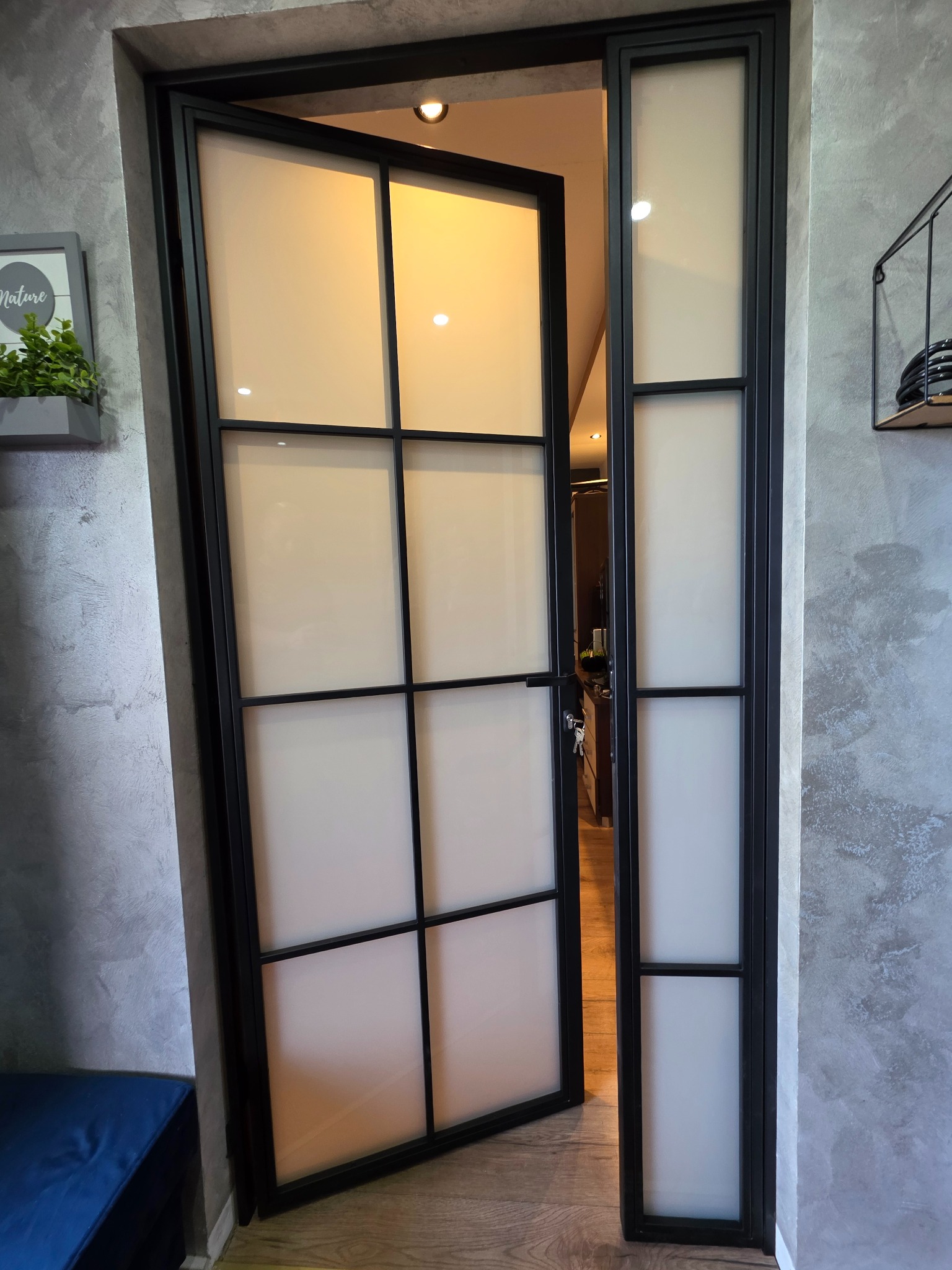 Otwarta, loftowa brama wewnętrzna z czarnymi profilami i mlecznym szkłem, widoczna z boku na tle szarej ściany i fragmentu podłogi.