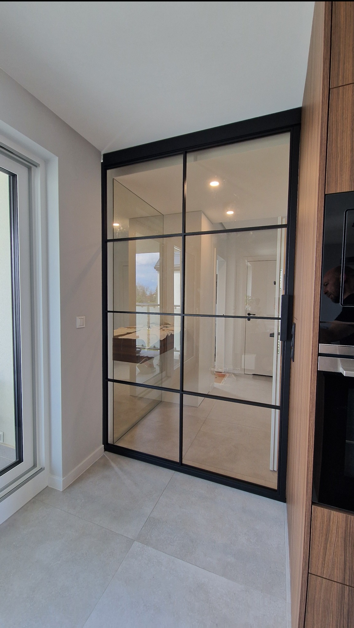 Nowoczesna, dwuskrzydłowa przeszklona ściana działowa z czarnymi szprosami w minimalistycznym wnętrzu. Widoczne odbicie osoby w szybie piekarnika.