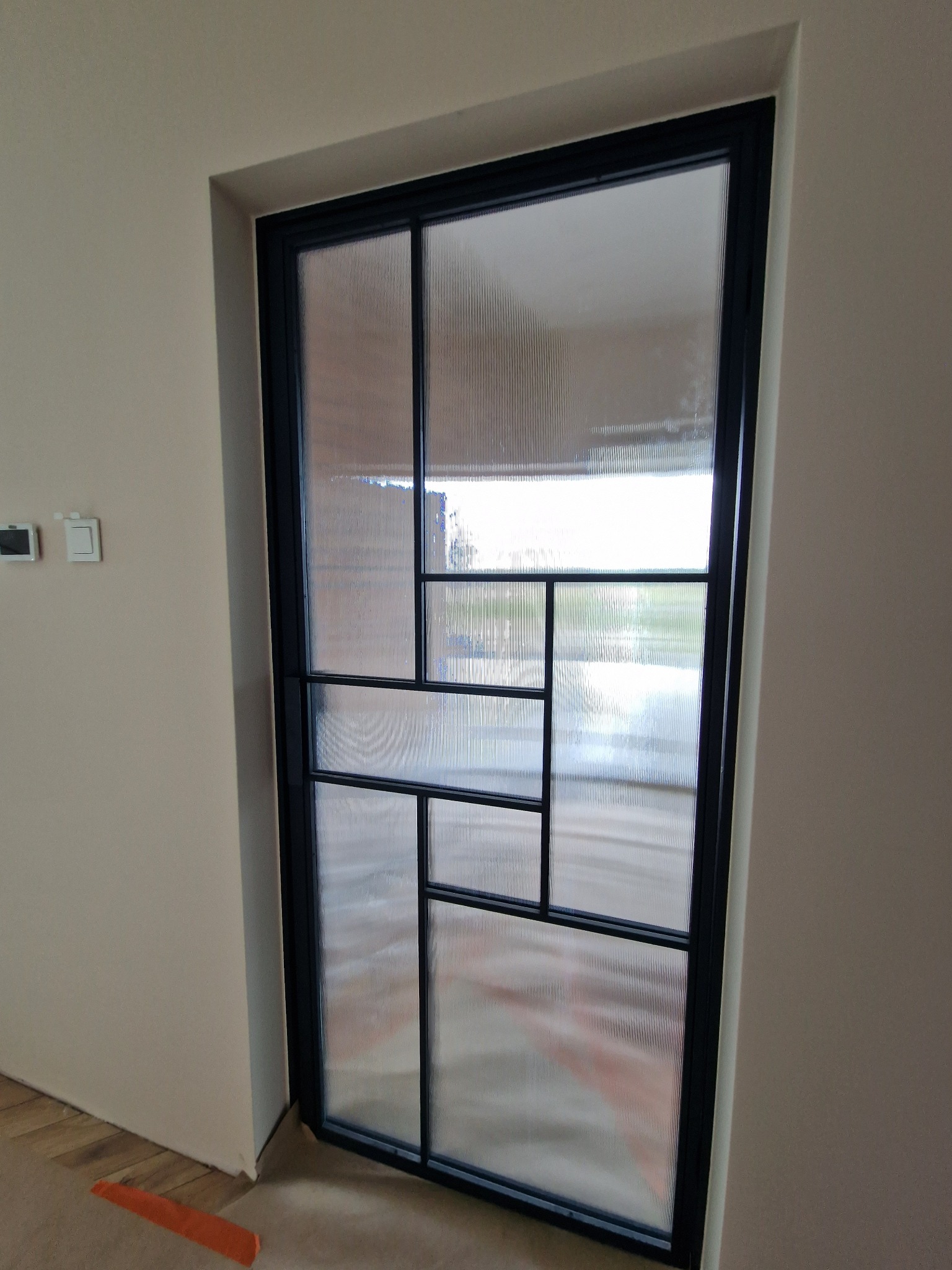 Nowoczesne drzwi wewnętrzne z czarną ramą i szkłem ornamentowym w geometryczne wzory, wkomponowane w jasną ścianę.