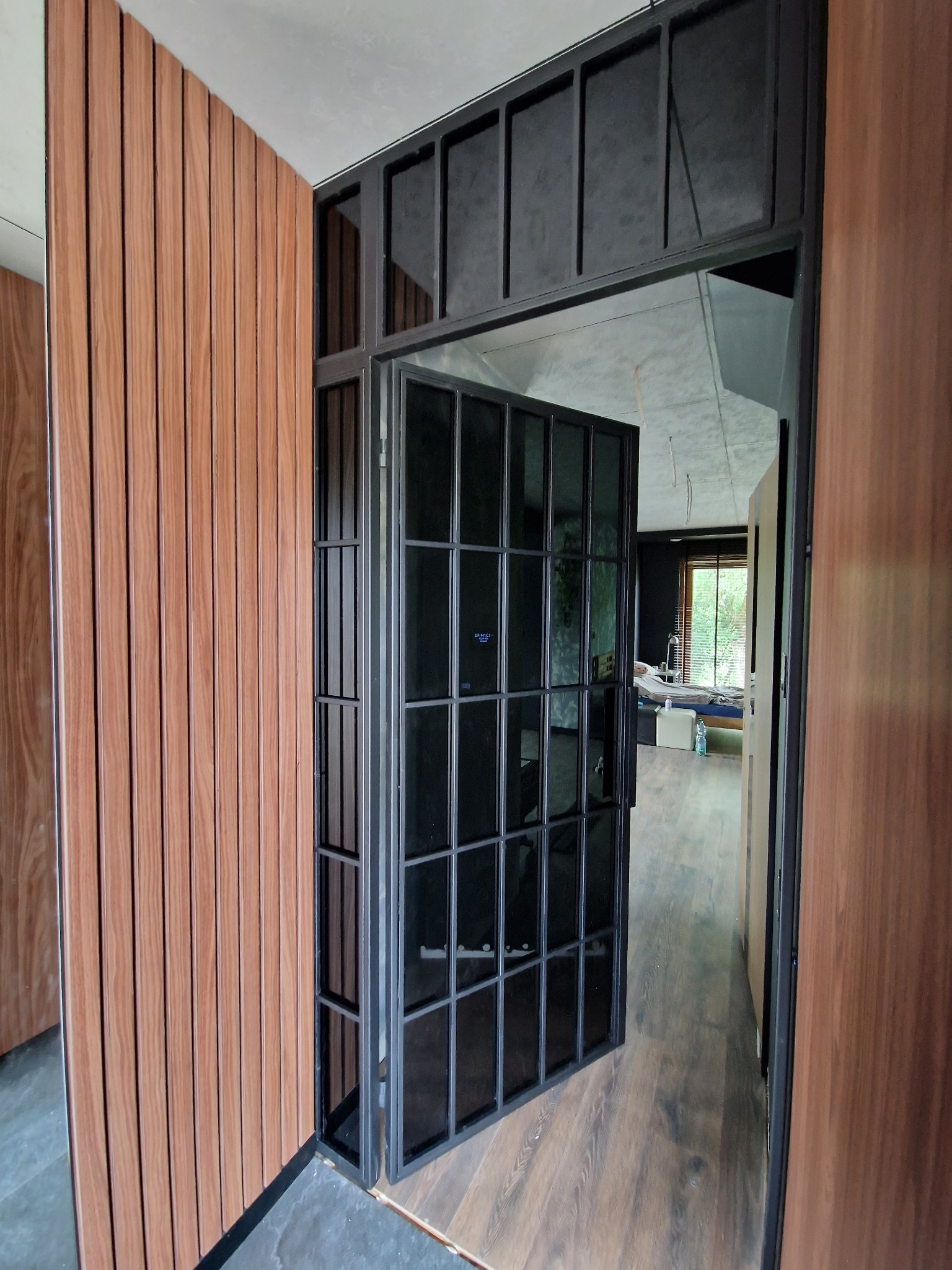 Nowoczesne, czarne, metalowe drzwi z szybami w loftowym stylu, otwarte do wnętrza. Obok widoczna drewniana okładzina ścienna w ciepłym odcieniu.