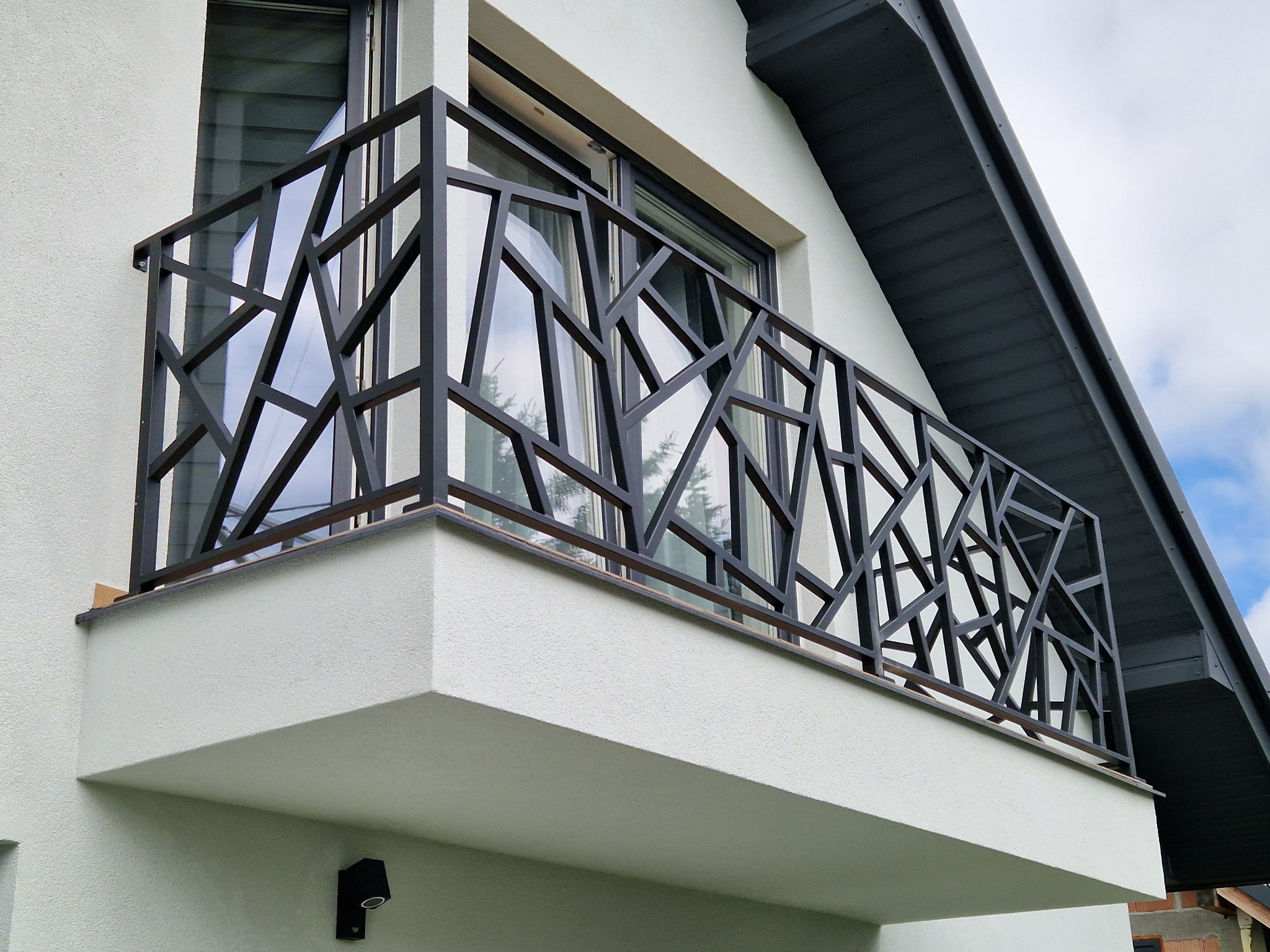 Nowoczesna, metalowa balustrada balkonowa z geometrycznym wzorem na tle białej elewacji domu z oknem. Balustrada w kolorze grafitowym.