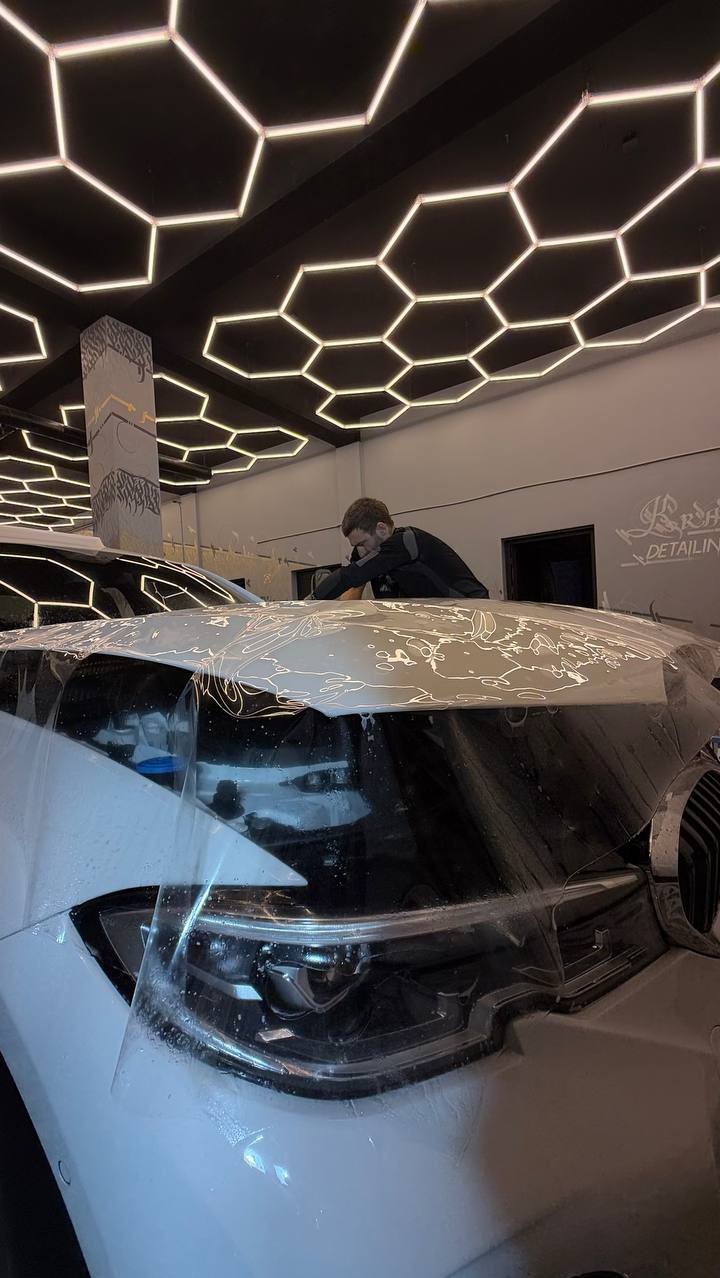 Pracownik aplikuje folię ochronną na maskę białego samochodu w jasnym wnętrzu studia detailingowego z heksagonalnym oświetleniem sufitowym.