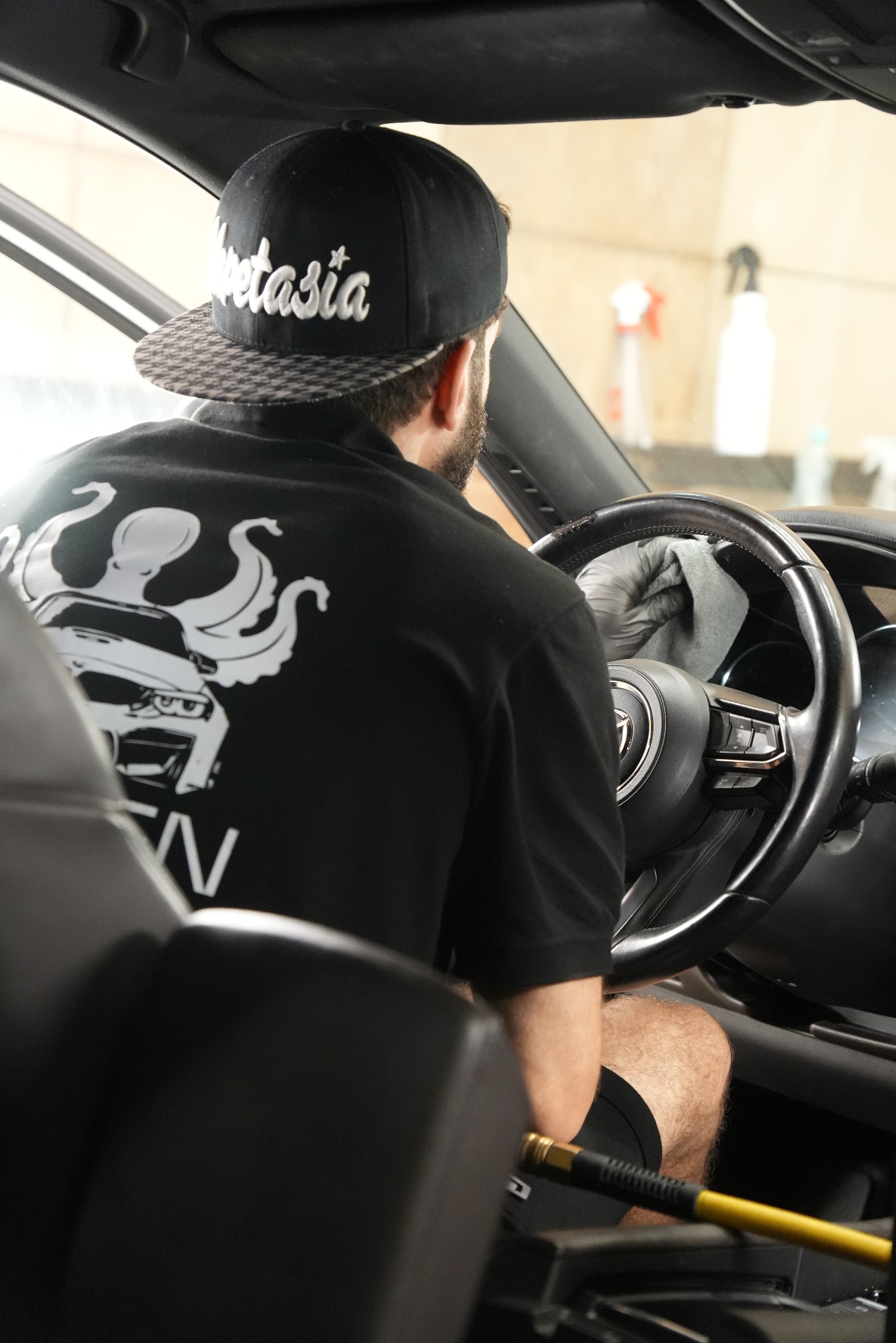 Wnętrze auta: detailer w czapce i rękawicach czyści kierownicę szarą ściereczką. W tle butelki ze środkami czyszczącymi i wąż do sprężonego powietrza.