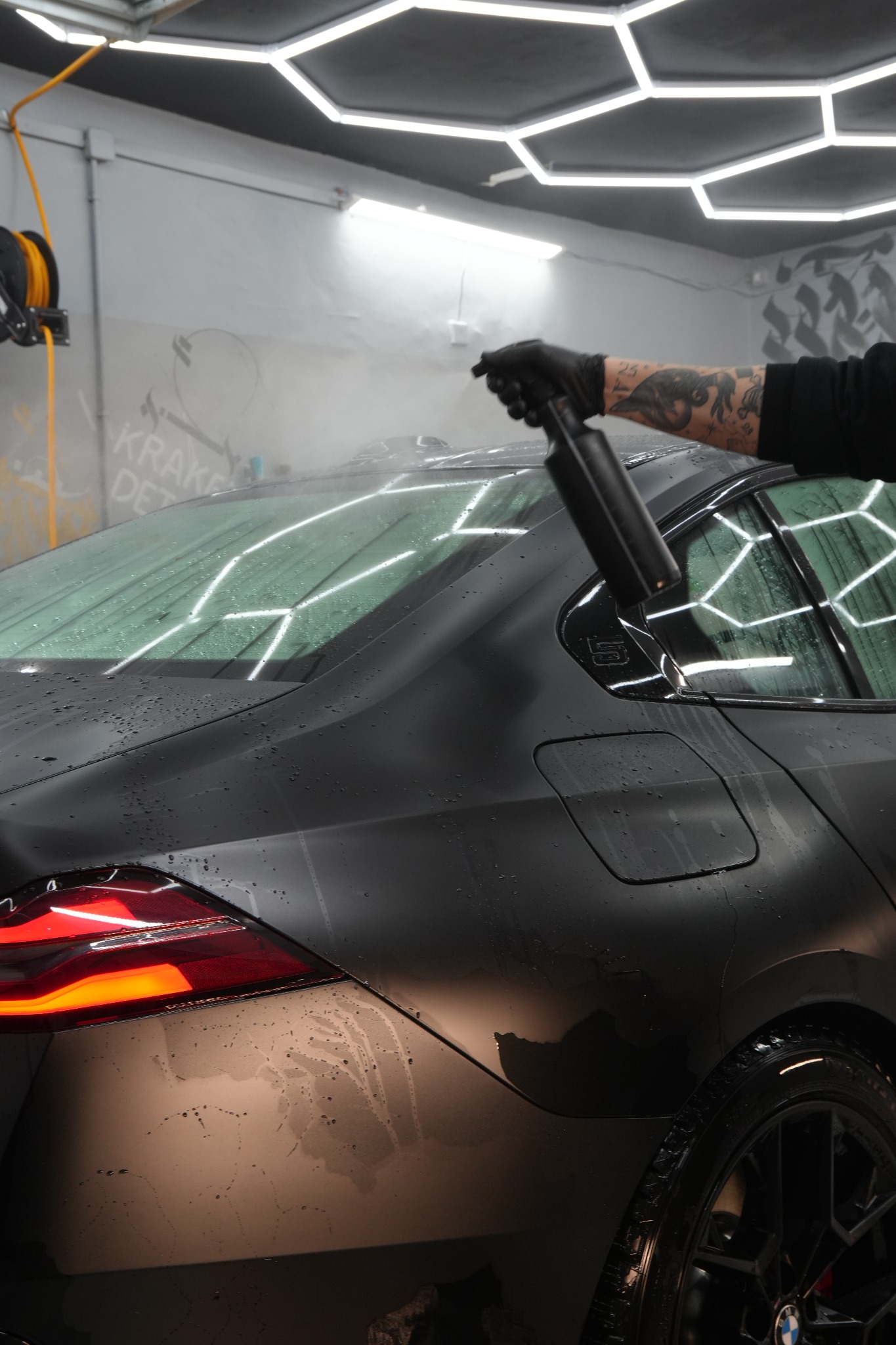 Matowy, czarny samochód w trakcie detailingu; widać spryskiwacz w dłoni osoby w czarnej rękawicy, oświetlenie heksagonalne, krople wody na karoserii.