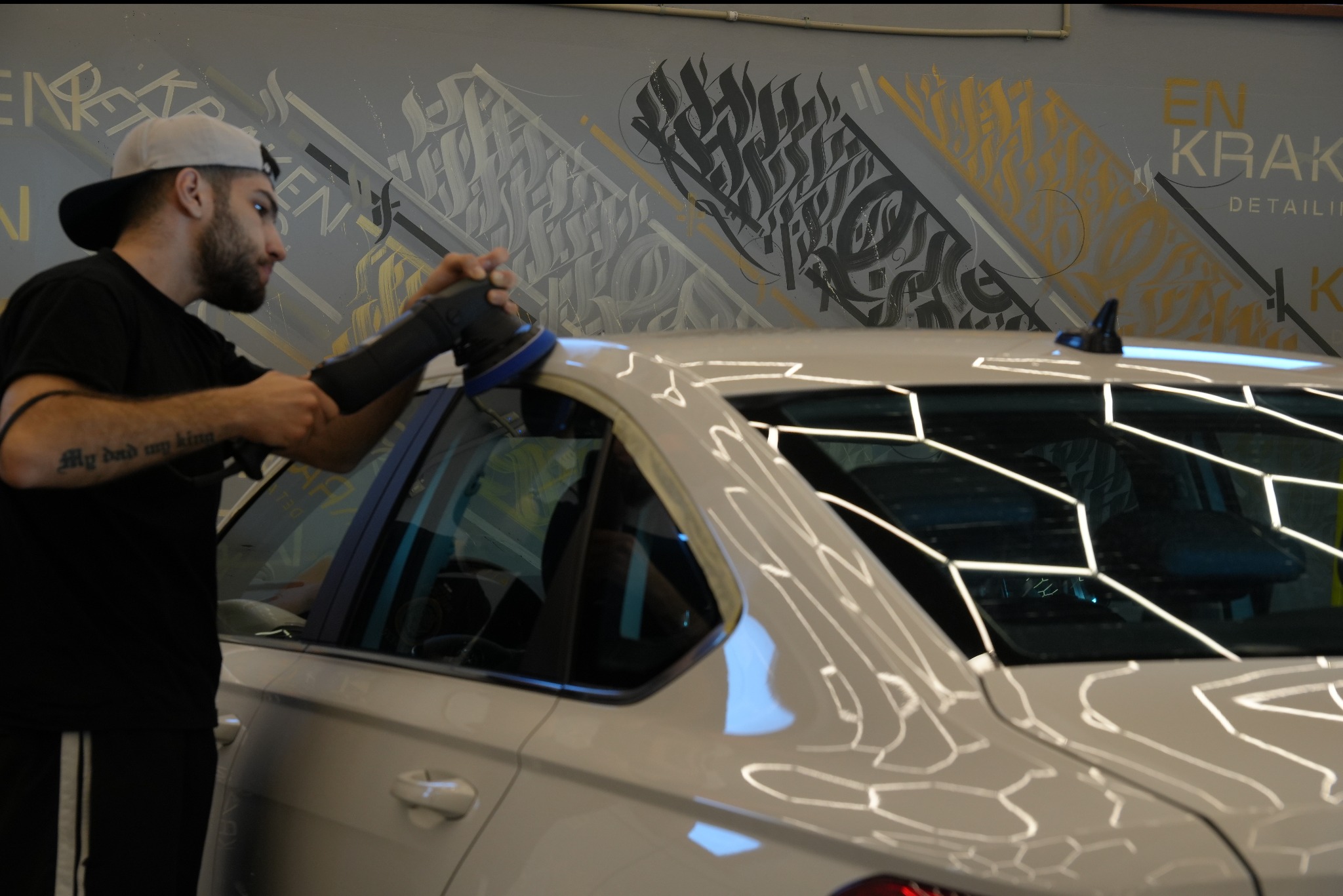 Detailing auta: mężczyzna poleruje biały samochód w studiu z graffiti na ścianie. Widoczny fragment karoserii i narzędzie polerskie.