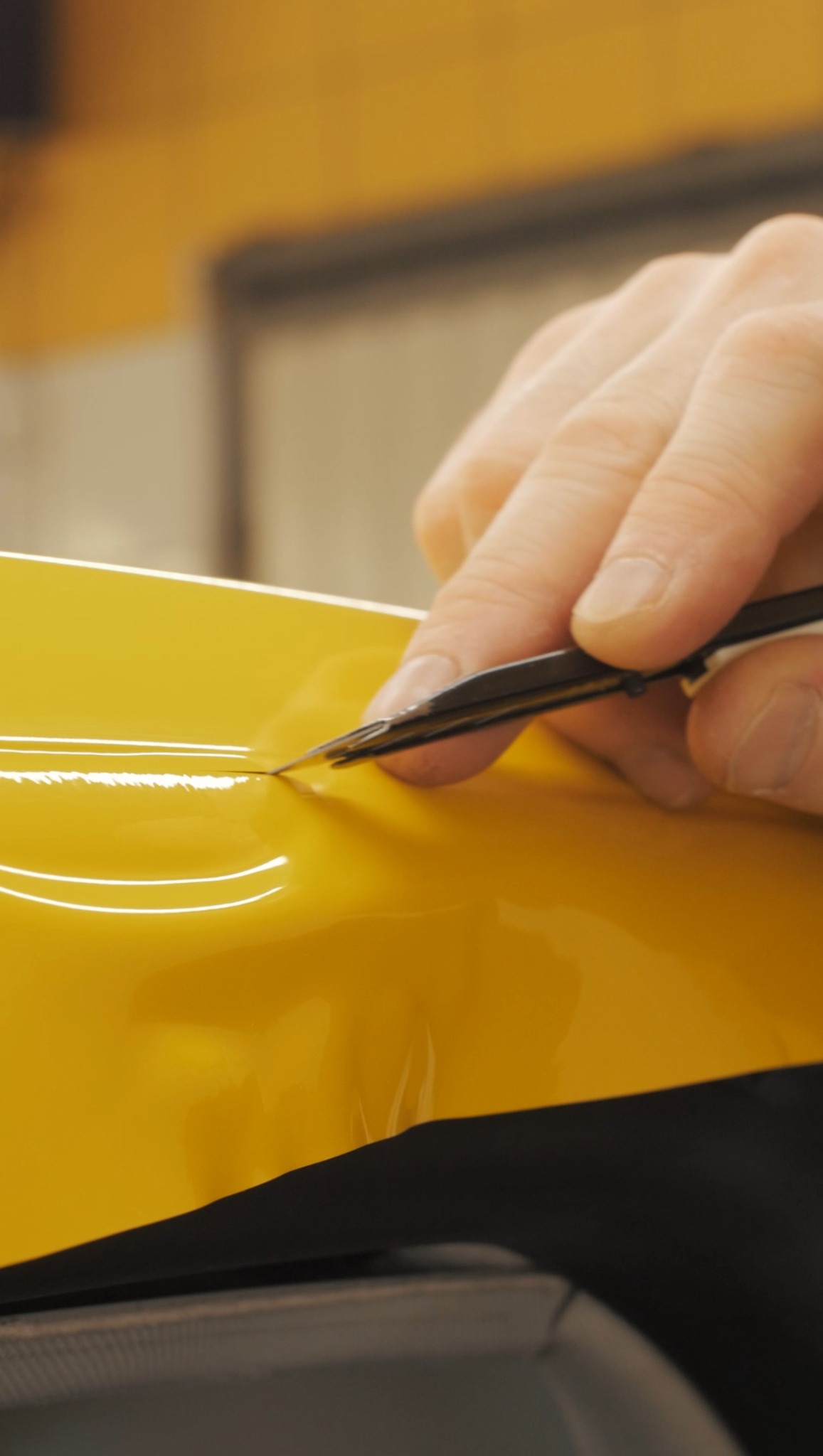 Precyzyjne docięcie żółtej folii na karoserii auta za pomocą pęsety podczas aplikacji, zbliżenie na detal, oświetlenie warsztatowe.