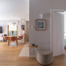 Adrian Tokarz Marketing - Jasne, przestronne wnętrze mieszkania z drewnianą podłogą, otwartymi drzwiami do sypialni i osobą ustawiającą krzesło przy okrągłym stole. Minimalistyczny design.