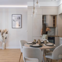 Adrian Tokarz Marketing - Jasne wnętrze: okrągły stół z nakryciem, krzesła, szafki kuchenne z otwartą szafką, kobieta sięga do szafki, dekoracje z suszonych traw, telewizor na ścianie.