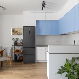 Adrian Tokarz Marketing - Jasne wnętrze kuchni z drewnianym stołem i szarymi krzesłami. W tle zabudowa kuchenna z błękitnymi szafkami i czarna lodówka. Na podłodze panele imitujące drewno.