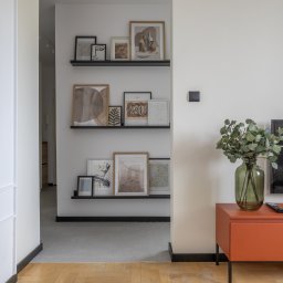 Adrian Tokarz Marketing - Jasne wnętrze: minimalistyczny salon z drewnianą podłogą, półkami z grafikami, pomarańczową komodą i telewizorem. Wazon z zielonymi gałązkami dodaje naturalnego akcentu.